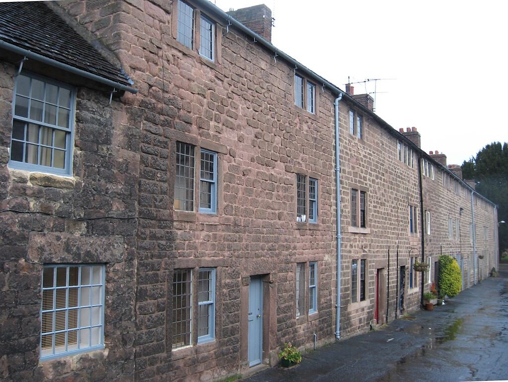 Fotografia przedstawia długi, dwukondygnacyjny, ceglany budynek z podwójnymi oknami i płaski dachem. Co jakiś czas widać drzwi wejściowe do budynku.