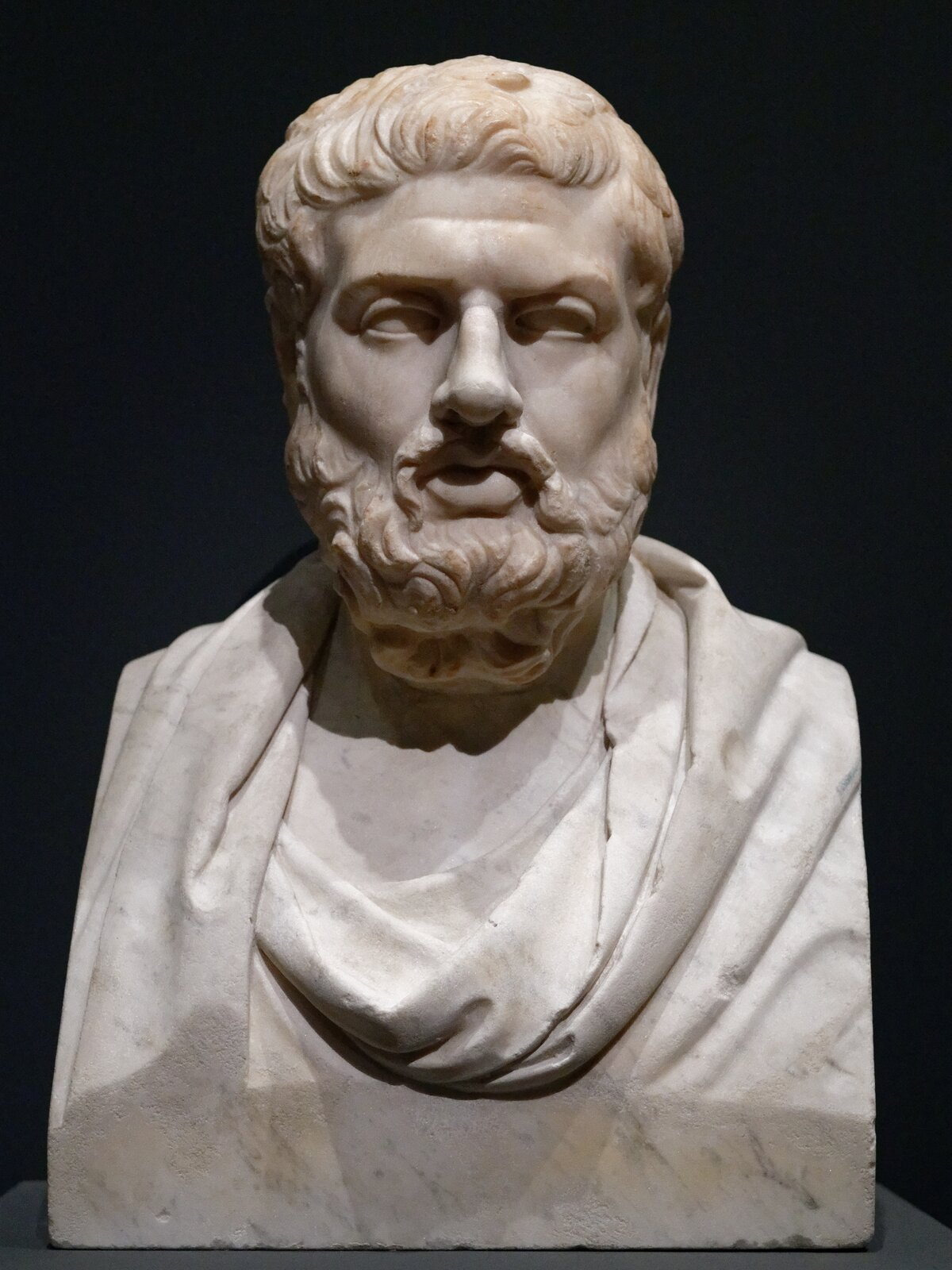 Ilustracja przedstawia rzeźbę „Teokryt” nieznanego autora. Rzeźba jest wykonana z jasnego kamienia. Przedstawia popiersie mężczyzny. Ma on kręcone włosy, wąsy i brodę. Na czole ma poziomą zmarszczkę. Dookoła szyi ma zarzuconą szatę. W tle jest czarna ściana. 