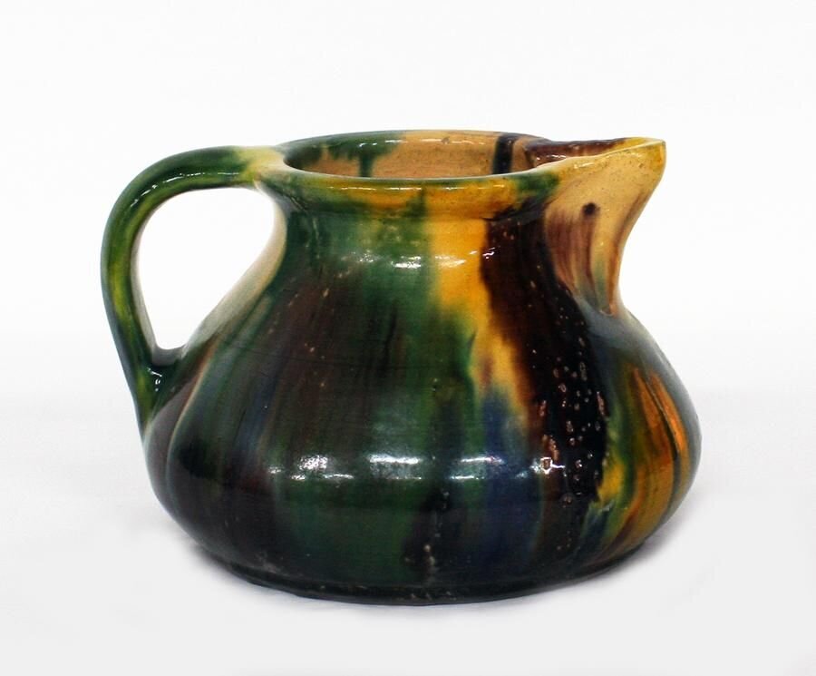 Fotografia przedstawia dzbanek szkliwiony. Dzbanek ma jeden uchwyt. Ma kolor ciemnozielony z pasmami brązu, beżu i żółci przy dzióbku. Dzban ma pękaty kształt. Jedno ucho znajduje się po lewej stronie, dziubek po prawej.