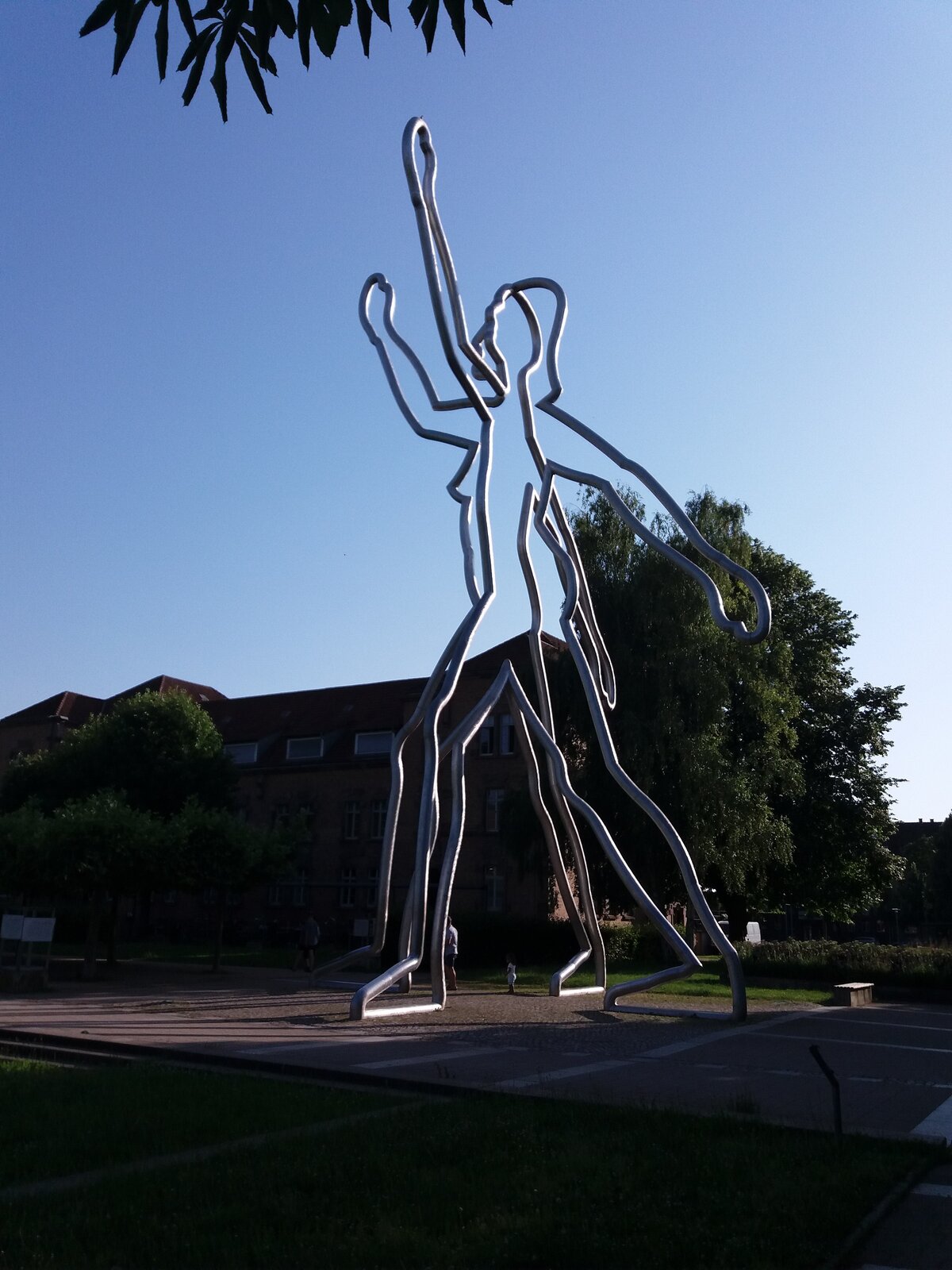 Fotografia przedstawia stojące duże figury konturowe zbudowane z rur, przypominające dwoje biegnących ludzi na jednym z placów wśród zabudowań. Wokół spacerują ludzie i rosną drzewa