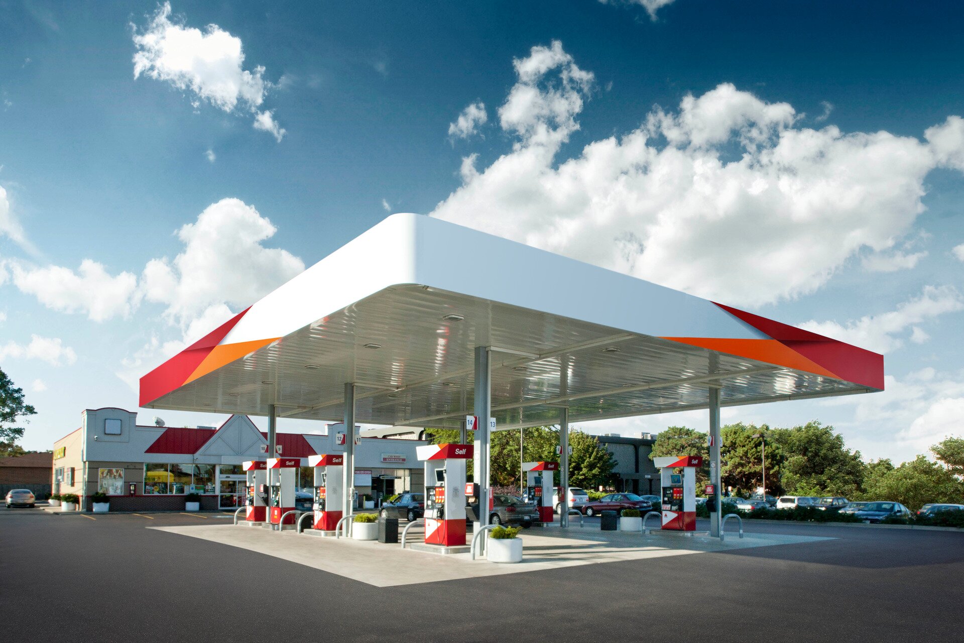 Ilustracja przedstawia budynek stacji benzynowej. Na stacji pod dachem znajdują się dystrybutory paliwa, za nimi znajduje się budynek ze sklepem. Po bokach stacji na parkingu stoją samochody.