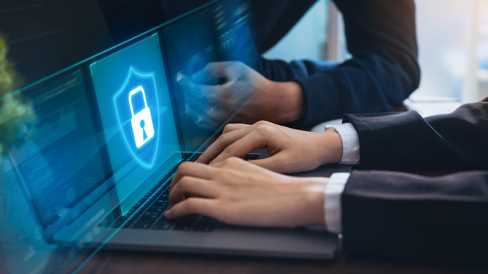 Zdjęcie przedstawia kobiece dłonie, które dotykają klawiatury laptopa. Na ekranie laptopa widoczna jest ikona tarczy z zamkniętą kłódką. W tle widoczna jest opierająca się o blat ręka.