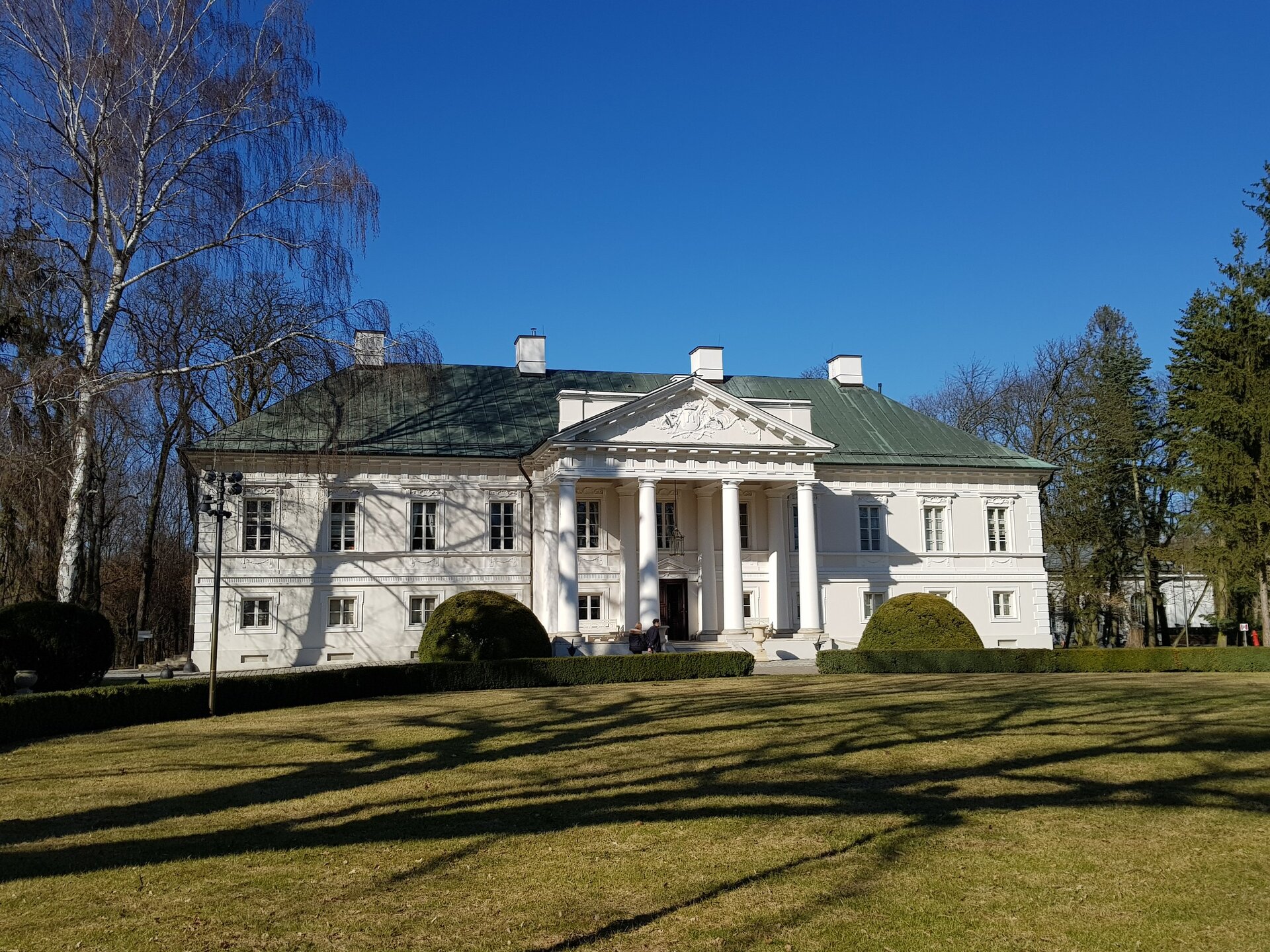 Zdjęcie przedstawia widok współczesny na pałac w Małej Wsi. W centralnej części portyk z czterema kolumnami i tympanonem na szczycie, osłaniającym fragment neorenesansowej attyki. Na szczycie czterospadowego dachu widoczne kominy. Fasada obiektu utrzymana w porządku klasycystycznym, z równomiernie rozłożonymi liniami okien w części parterowej i na piętrze. Przed portykiem widoczne dwa klomby, flankujące wejście do obiektu. Po lewej stronie widoczna bezlistna brzoza, po prawej stronie drzewa iglaste - jodły. W tle za nimi fragment zabudowy budynku pałacowego. Na pierwszym planie oświetlony światłem słonecznym trawnik, na który pada cień drzewa. Niebo bezchmurne.