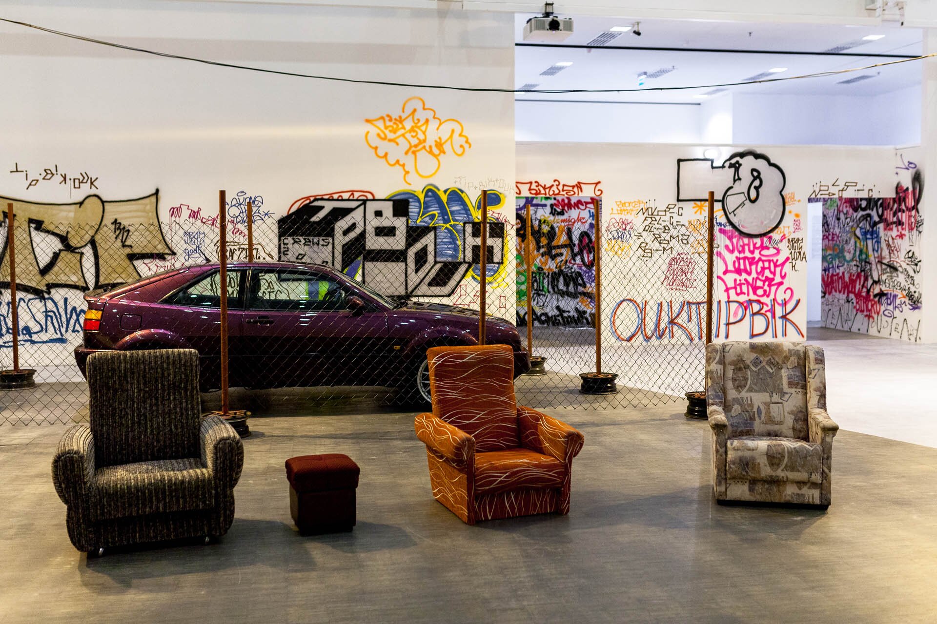 Fotografia Rafała Wyrwicha przedstawia salę z fragmentem ekspozycji pod tytułem Zajawka. Na pierwszym planie uchwycono trzy fotele – ciemnozielony z żółtawymi pionowymi liniami, pomarańczowy z jaśniejszymi, falowanymi liniami, oraz beżowy z brązowo‑szarymi wzorami o wielorakich kształtach. Pomiędzy dwoma pierwszymi fotelami stoi bordowy puf. Podłoga jest szara. Za fotelami jest ogrodzenie, za którym znajduje się fioletowy samochód Volkswagen Corrado oraz ściany pokryte różnokolorowym graffiti. 