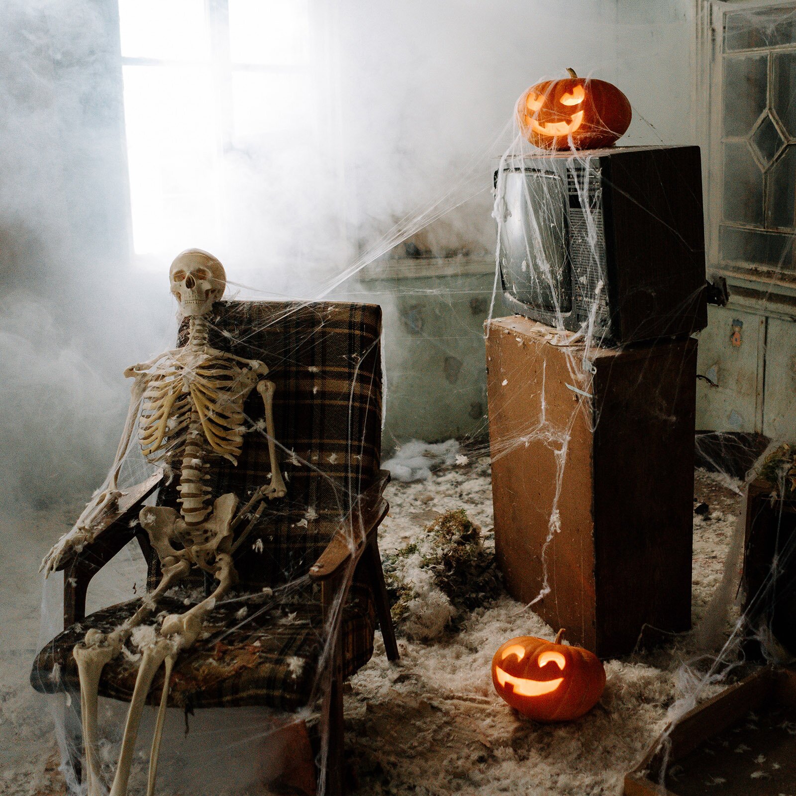 Zdjęcie przedstawia wnętrze starego pokoju. Na fotelu siedzi kościotrup obok niego widoczny stary telewizor kineskopowy otoczony pajęczyną oraz dwie wydrążone dynie, do których wstawiono świece. 