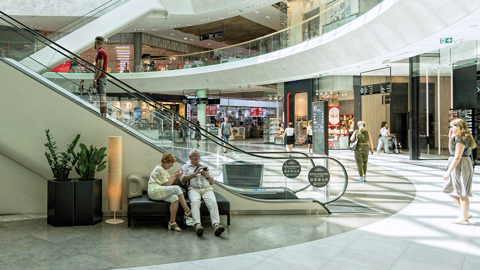 Zdjęcie przedstawia wnętrze galerii handlowej. Widoczne są schody ruchome prowadzące na piętro galerii. W tle znajdują się witryny sklepów. Na zdjęciu widoczni są także ludzie. Kobieta i mężczyzna siedzą na ławce przy schodach, ktoś wjeżdża po schodach.