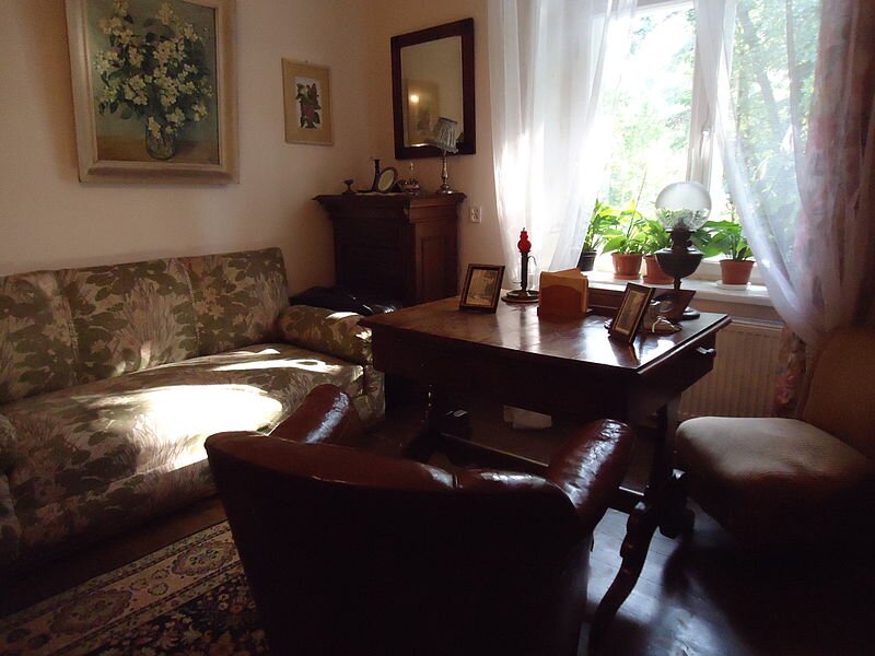 Kliknij, aby powiększyć Zdjęcie przedstawia wnętrze pokoju, w którym pod ścianą na wprost znajduje się kanapa, nad nią wiszą dwa obrazy. Na prostopadłej ścianie z dużym jasnym oknem wisi lustro i stoi wąska wysoka szafka. Na środku drewniane prostokątne biurko, na blacie ustawione są ramki ze zdjęciami. Do biurka dosunięty jest skórzany fotel.