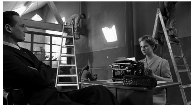 Zdjęcie przedstawia wnętrze fabryki. Robotnicy malują ściany. W sali stoi biurko, przy którym siedzi przy maszynie do pisania młoda kobieta o blond włosach. Naprzeciw niej siedzi Schindler.