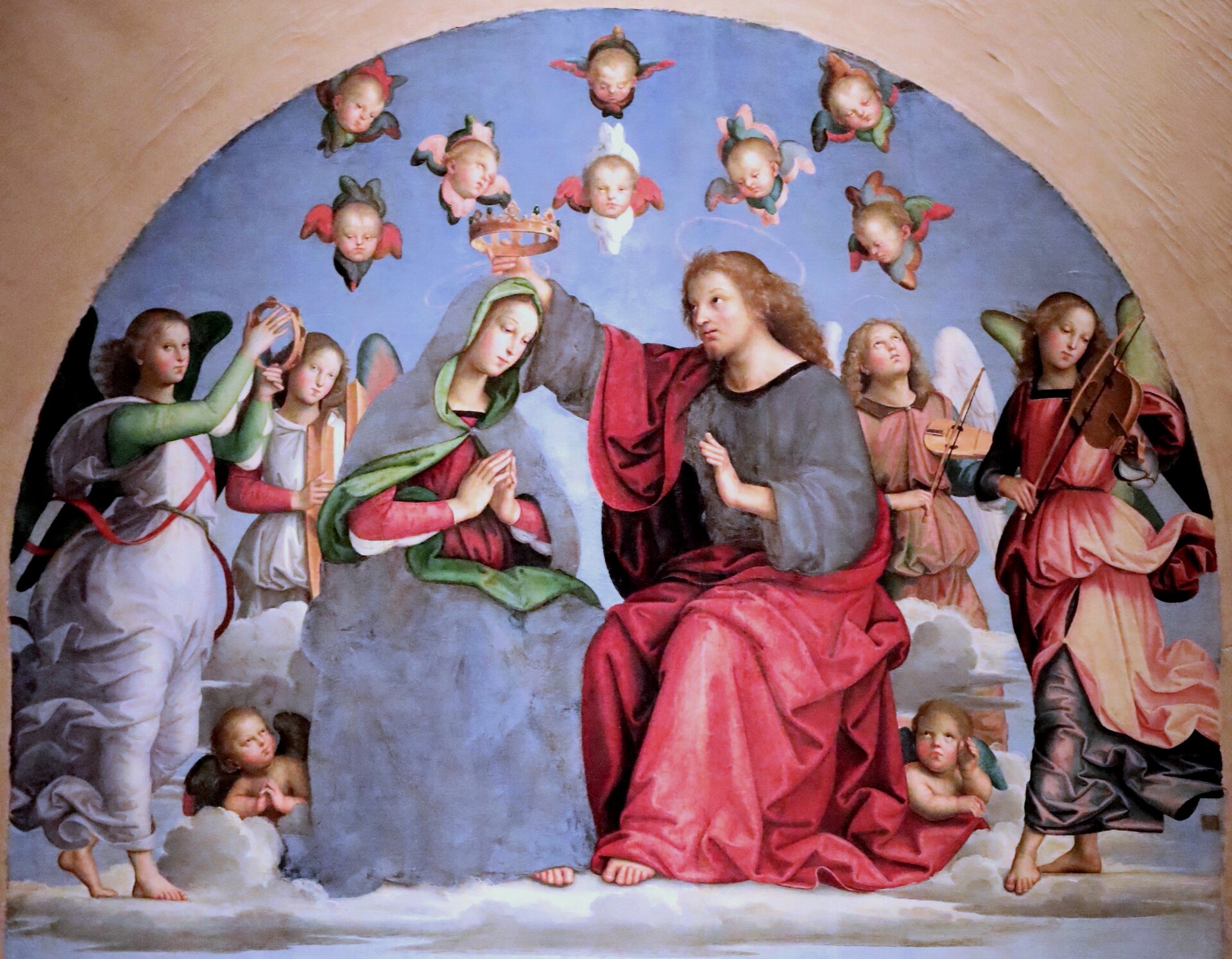 Ilustracja interaktywna przedstawia temperę „Koronacja Matki Bożej” autorstwa Rafaela Santi. Zamknięta w łuku kompozycja ukazuje siedzące postacie Chrystusa i Matki Bożej wśród aniołów. Młody mężczyzna o jasnych, długich włosach, ubrany w szarą szatę, okrytą czerwonym płaszczem w wyciągniętej nad głową Marii dłoni trzyma koronę. Kobieta, o delikatnych rysach twarzy i złożonych do modlitwy dłoniach, schyla głowę w kierunku Jezusa. Matka Boża ubrana jest w czerwoną suknię, którą szczelnie okrywa wraz z włosami szary, podbity zielenią płaszcz. Na około pary artysta umieścił grających na instrumentach aniołów. Skrzydlate postacie ubrane są w powłóczyste, przypasane w talii, różnobarwne stroje. Przy nogach Marii i Jezusa siedzą małe, skrzydlate putta. Z jasno-niebieskiego nieba wychylają się głowy małych aniołków. Namalowane postacie unoszą się na jasnych obłoczkach.