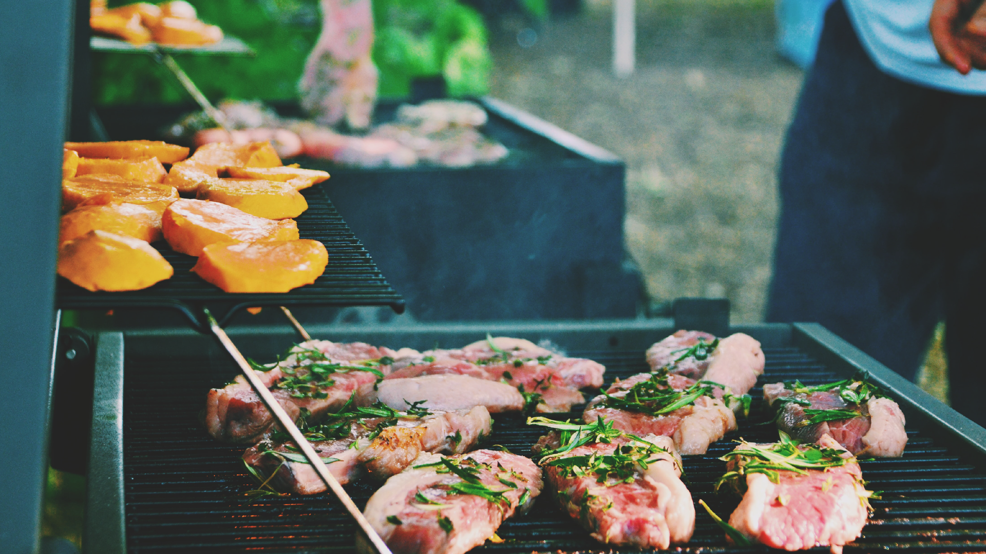 Zdjęcie na pierwszym planie przedstawia grill, na którym ułożone są kawałki mięsa przybrane świeżymi przyprawami.. Na dalszym planie widoczne jest kolejny grill, na którym ułożone są inne produkty.