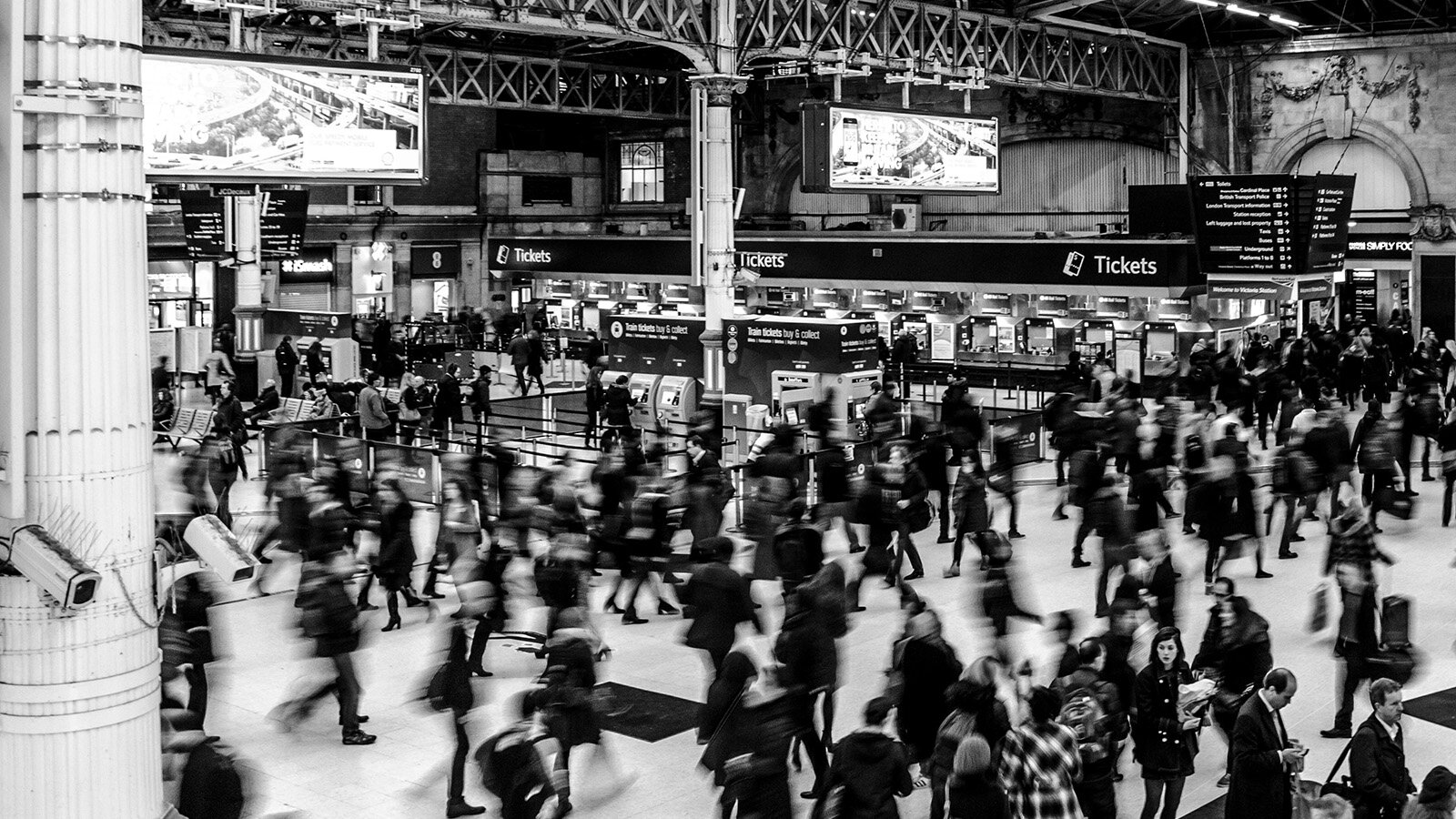 Czarno-biała fotografia przedstawia halę dworca kolejowego w stylu industrialnym, wypełnioną ludźmi przemieszczającymi się w różnych kierunkach. Postacie poruszają się szybko i są rozmyte. Widoczny jest długi rząd automatycznych kas sprzedaży biletów. Nad kasami widoczny jest powtarzający się napis w języku angielskim: TICKETS. Przed nimi widoczne są dwa stanowiska sprzedaży biletów, nad którymi widnieje napis w języku angielskim: Train tickets buy 6 collect. U góry widoczne są duże ekrany, na których wyświetlana jest linia kolejowa. Widoczne są również znaki informacyjne.