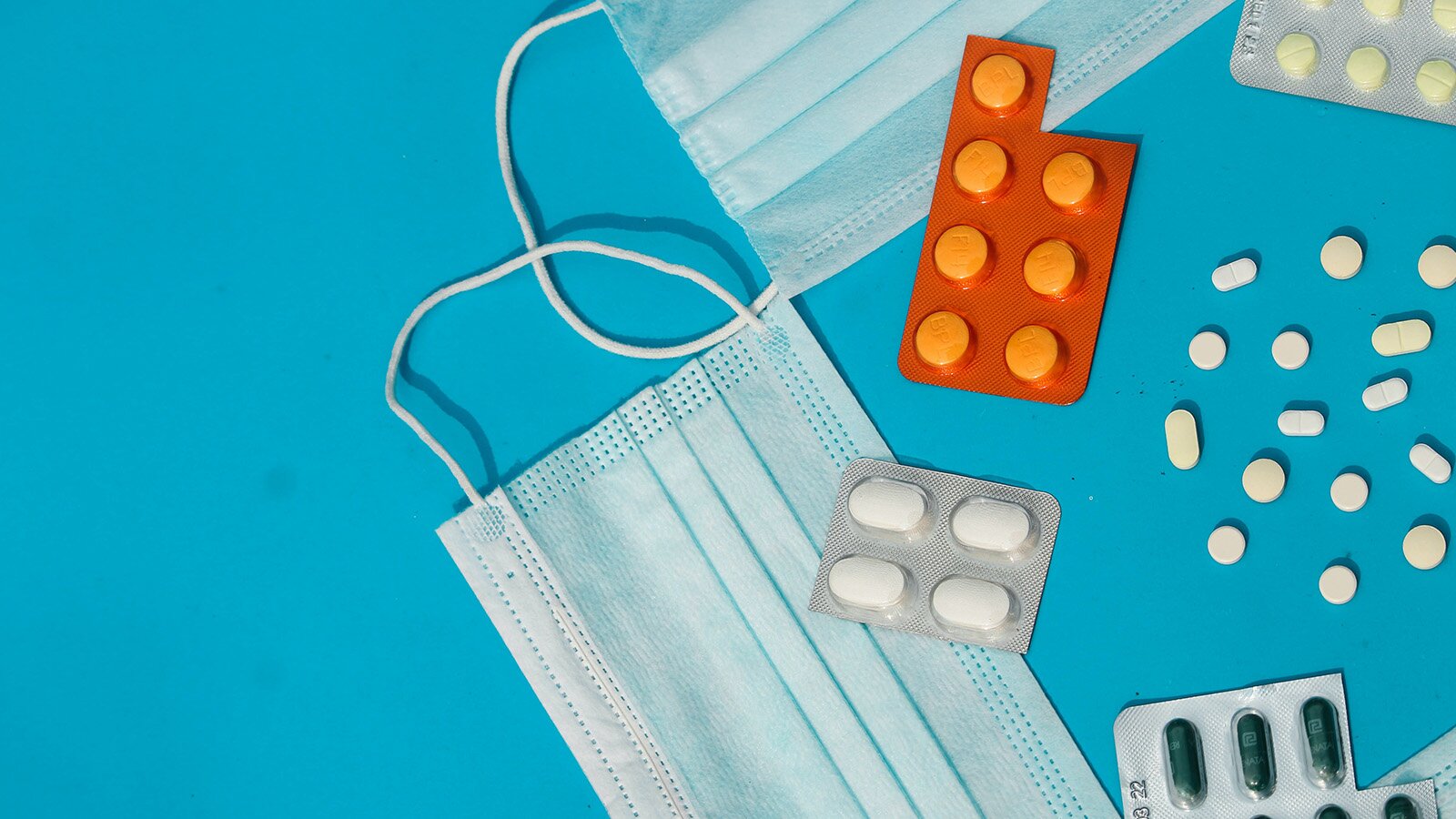 Zdjęcie przedstawia ułożone na blacie blistry tabletek. Kilka z nich leży rozsypanych. Widoczne są również dwie maseczki ochronne. 