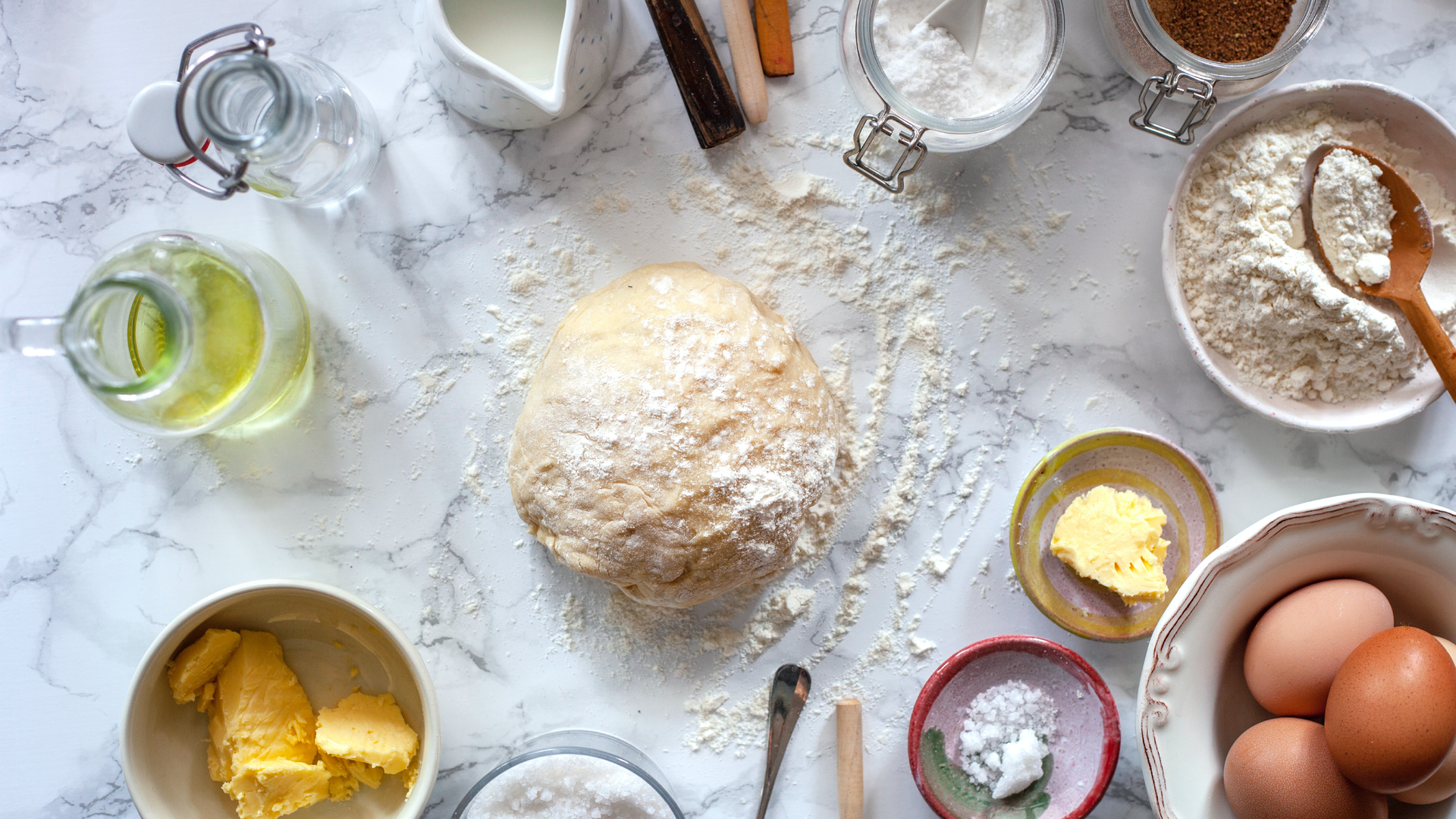 Na zdjęciu widoczne są składniki potrzebne do upieczenia ciasta. Na środku kuchennego blatu leży kulka surowego ciasta, posypana mąką. Wokół znajdują się: woda, olej, mleko, masło, sól, mąka, jajka.