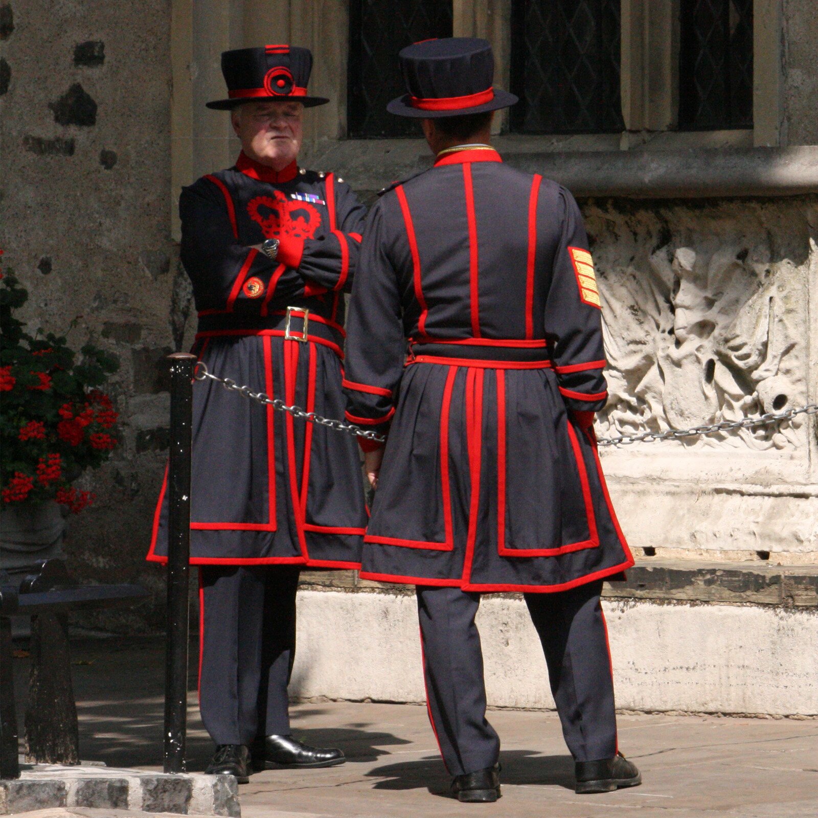 Zdjęcie przedstawiające dwóch mężczyzn ubranych w czarne stroje z czerwonymi elementami oraz kapelusze w tych samych kolorach. Mężczyźni stoją naprzeciw siebie, jeden z nich ma ręce założone na piersi. Stoją przed budynkiem.
