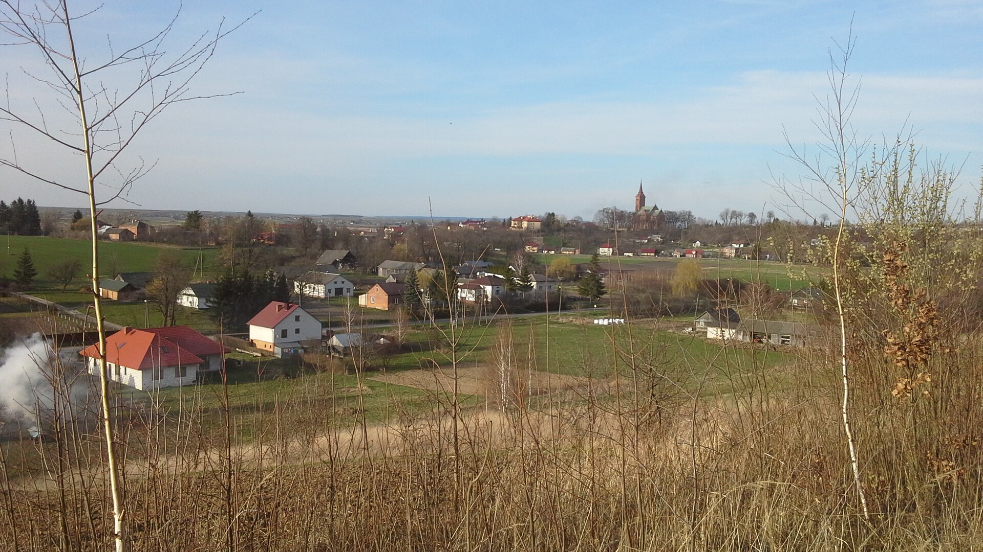 Fotografia wykonana z lekkiego wzniesienia ukazująca niewielką osadę złożoną z domów jednorodzinnych między polami uprawnymi i lasami.
