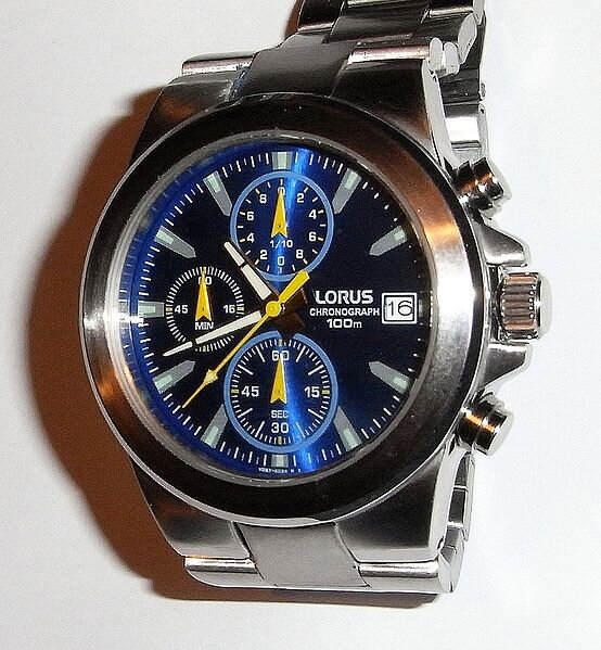 Fotografia przedstawia ręczny zegar kwarcowy. Na białym tle widać czarny okrąg z naniesionymi na brzegach srebrnymi krótkimi odcinkami, dłuższe określają godziny, krótsze‑minuty. W środku zaczepione cienkie wskazówki: białe‑godzinowa i minutowa oraz żółta‑sekundowa. Na czarnym tle widać również trzy mniejsze jasne okręgi z cyframi arabskimi na brzegach o czarnym tle z żółtymi wskazówkami strzałkami oraz napis firmy z okienkiem i cyfrą szesnaście.  Całość umieszczona w srebrnej obudowie, która z prawej strony posiada trzy srebrne wypustki w kształcie małych walców (pokrętła). Do obudowy doczepiony jest metalowy pasek składający się z metalowych prostokątów.