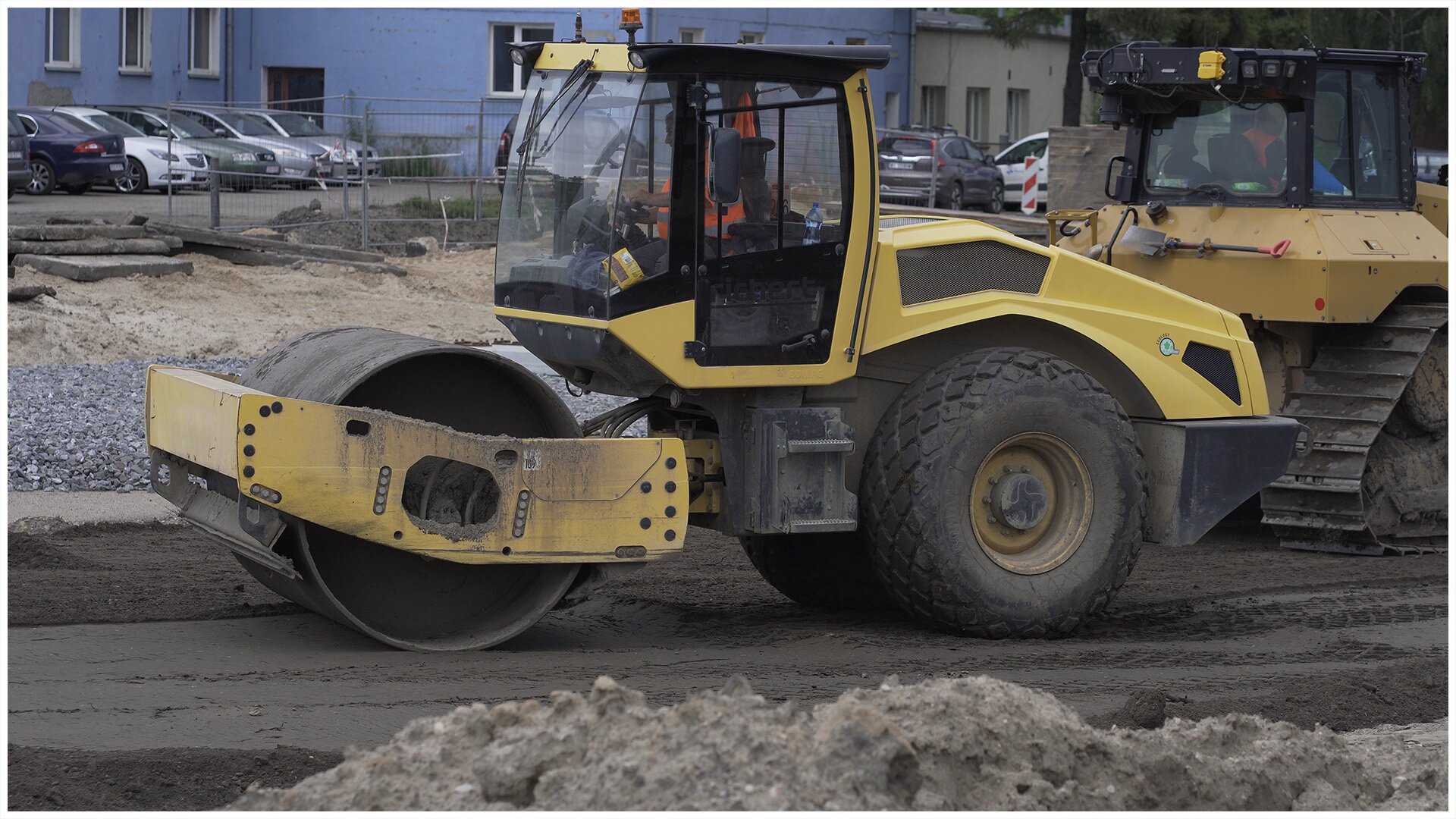 Ilustracja przedstawia walec drogowy. Tylna oś jest osadzona na kołach drogowych, a z przodu pojazdu znajduje się walec, którego zadaniem jest zagęszczanie kruszyw lub gruntów.