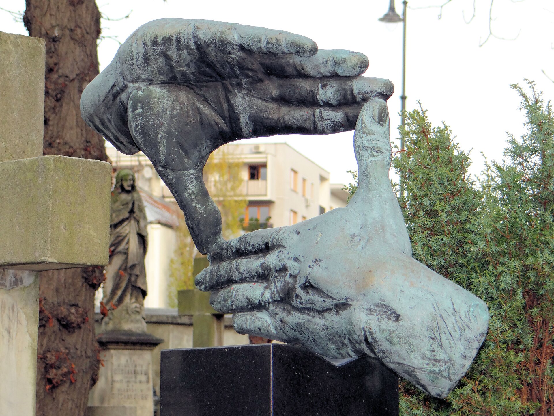 Zdjęcie przedstawia rzeźbę znajdującą się na nagrobku. To złożone dwie dłonie w taki sposób, że między nimi powstaje prostokąt naśladujący widok okiem kamery.