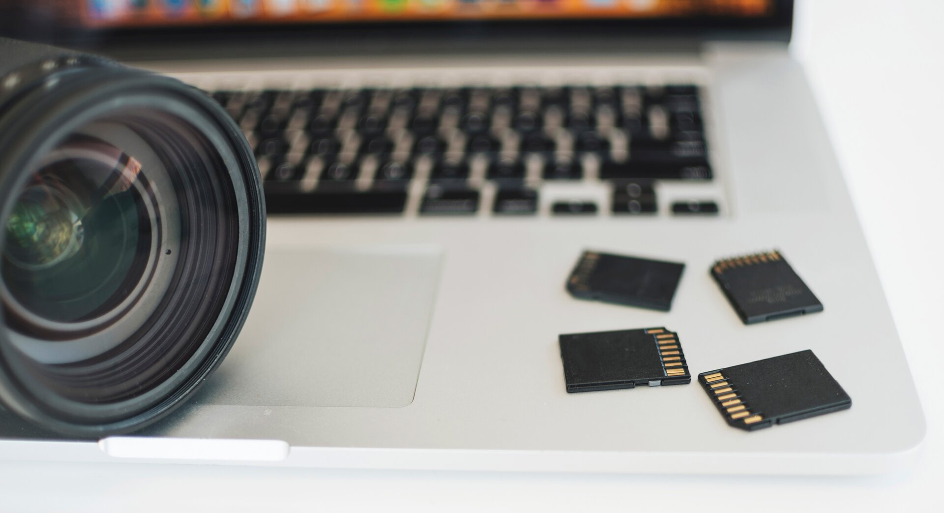 Zdjęcie przedstawia fragment białego blatu, na którym widoczne są po lewej stronie – obiektyw aparatu, po prawej stronie – cztery prostokątne karty pamięci do aparatu. W tle, widoczna rozmazana czarna klawiatura. 