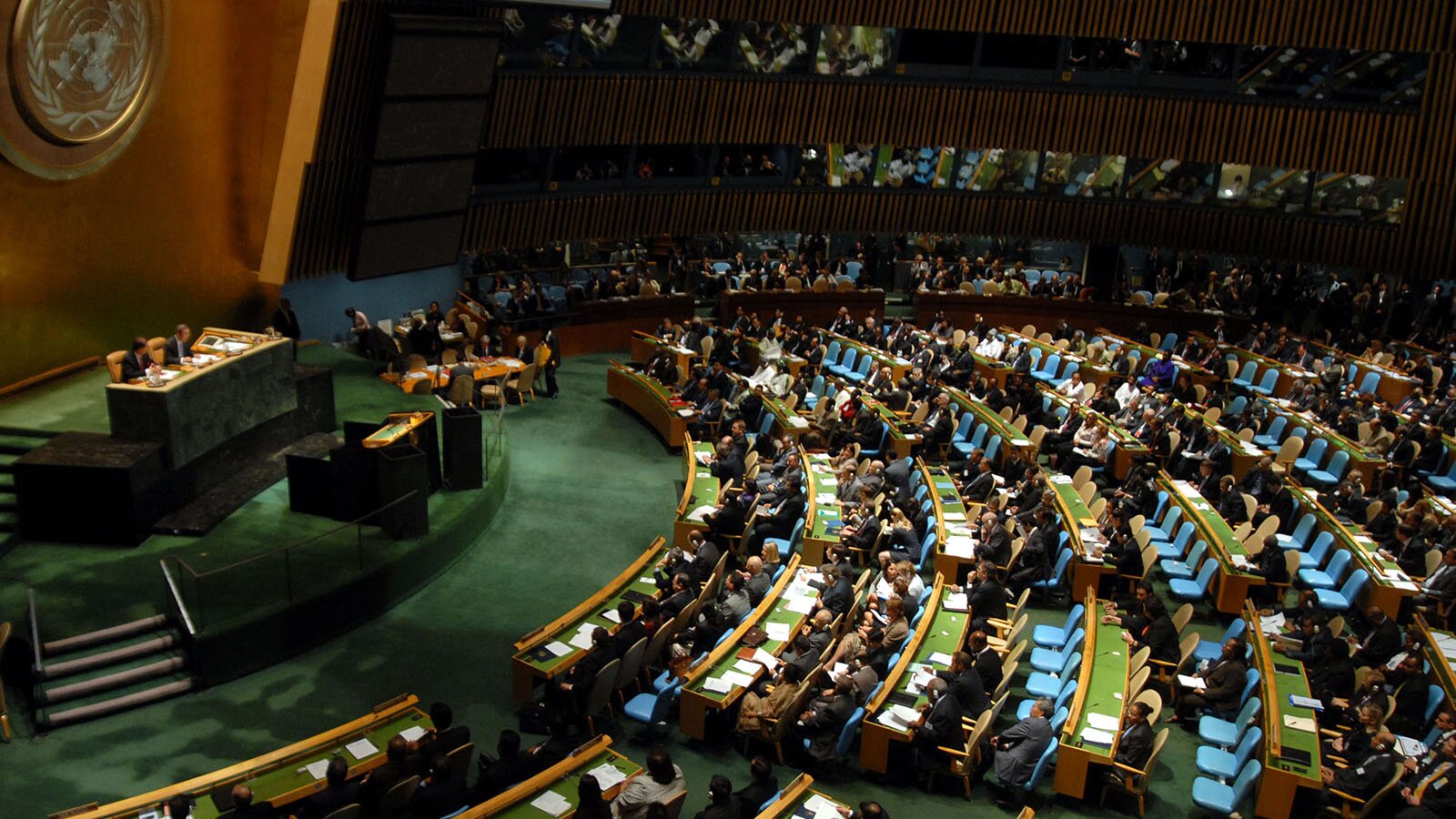 Zdjęcie przedstawia dużą aulę. Po lewej stronie widoczne jest podium, w tle którego widoczne jest godło Narodów Zjednoczonych. Na podium widoczne, u góry – duży stół, a poniżej – mównica. Pośrodku i po prawej stronie widoczne są rzędy niebieskich foteli ze stolikami. Na wielu fotelach siedzą ludzie.