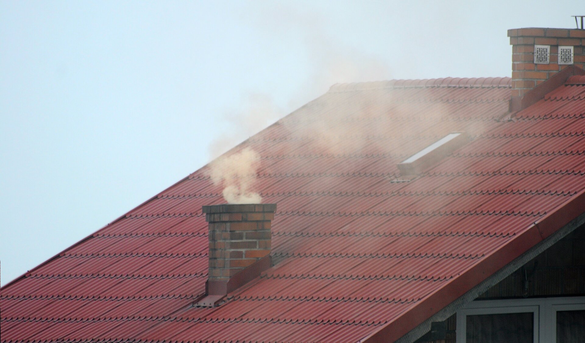 Na zdjęciu jest fragment dachu z kominem. Z komina wydobywa się żółty dym. 