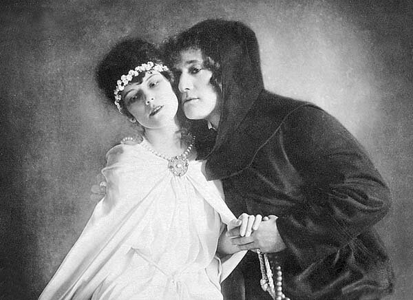 Czarno‑białe zdjęcie przedstawia przytuloną policzkami parę młodych ludzi. Od lewej strony młoda kobieta w białej sukni z białym wiankiem na głowie, przytula się lewym bokiem do mężczyzny w czarnym stroju z kapturem na głowie. Mężczyzna trzyma kobietę za lewą rękę. Oboje patrzą w dal. Tło jest szare.