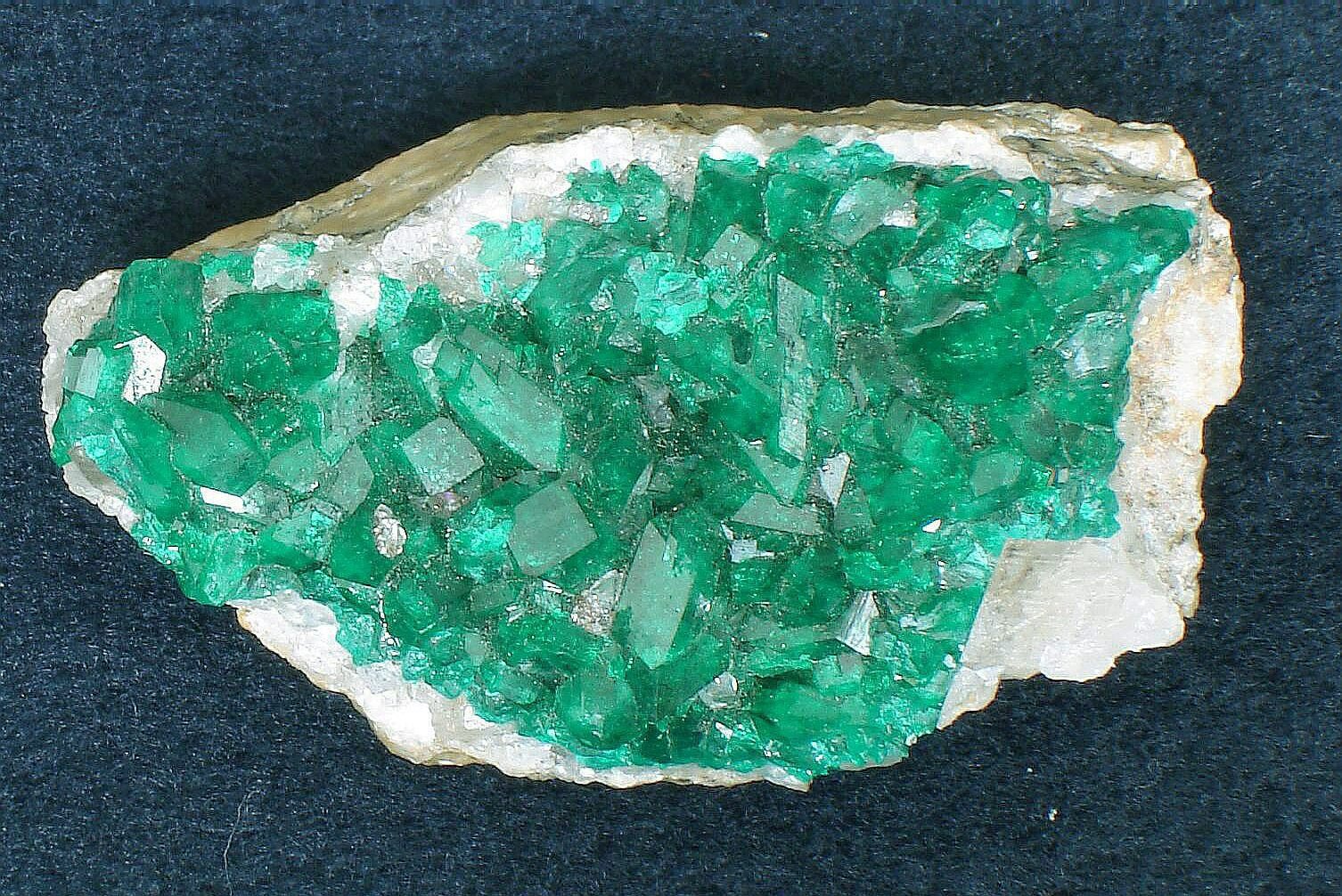 Zdjęcie ukazuje zielone, drobne kryształy. Mają zróżnicowane kształty. Znajdują się na powierzchni białego kamienia.  