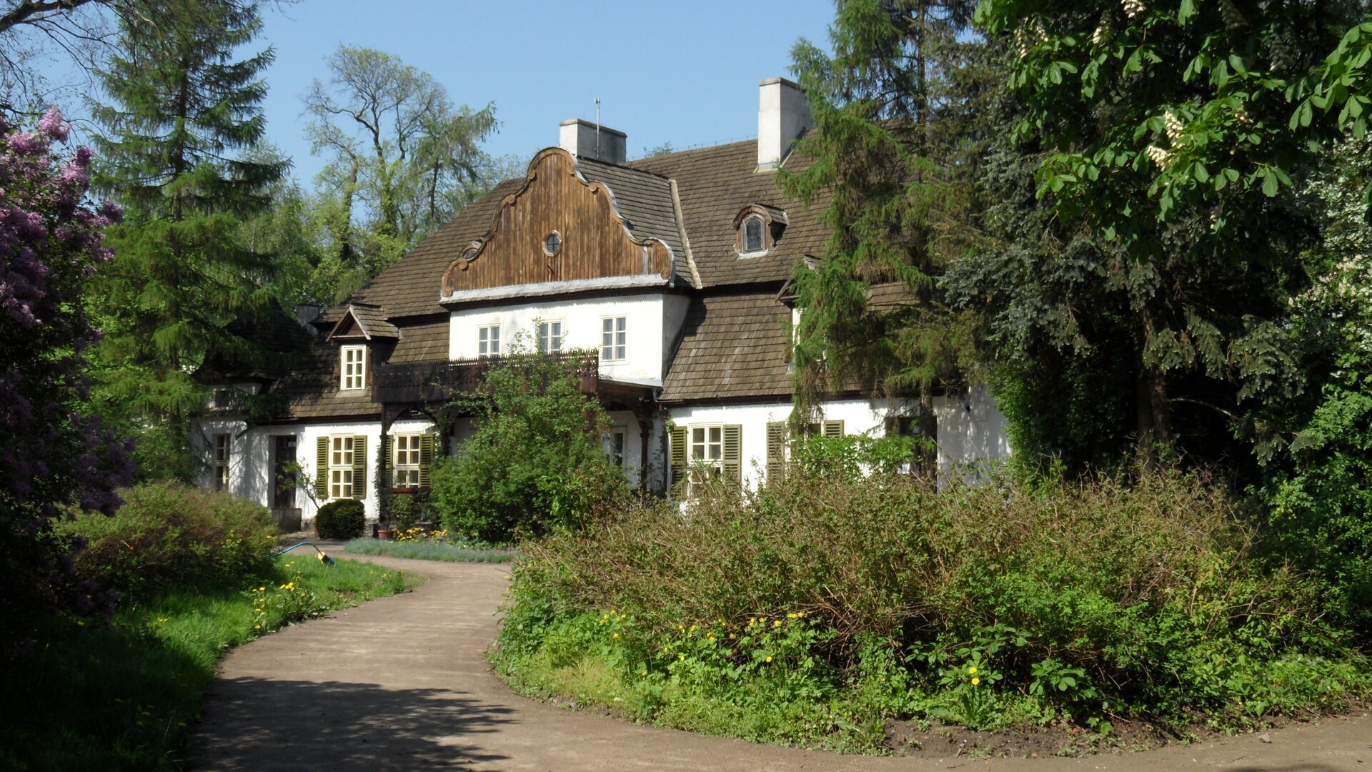 Fotografia przedstawia pobielony dworek szlachecki, otoczony krzewami i wysokimi drzewami. Jest to budynek parterowy, z rozbudowanym poddaszem. Dom pokryto gontem. Na pierwszym planie znajduje się okrągły klomb z podjazdem. Przed wejściem, pośrodku budynku, znajduje się ganek porośnięty dzikim winem, które osłania wejście do budynku. Nad gankiem znajduje się balkon z drewnianymi barierkami. Parterowe, prostokątne okna opatrzone są drewnianymi okiennicami. Na poddaszu, nad gankiem są trzy okna, na krańcach dachu, po jednej i drugiej stronie umieszczono po jednym małym, zadaszonym oknie. U samego szczytu dachu znajdują kolejne dwa małe, półokrągłe okna. Nad dworem górują dwa kominy. W tle bezchmurne niebo.
