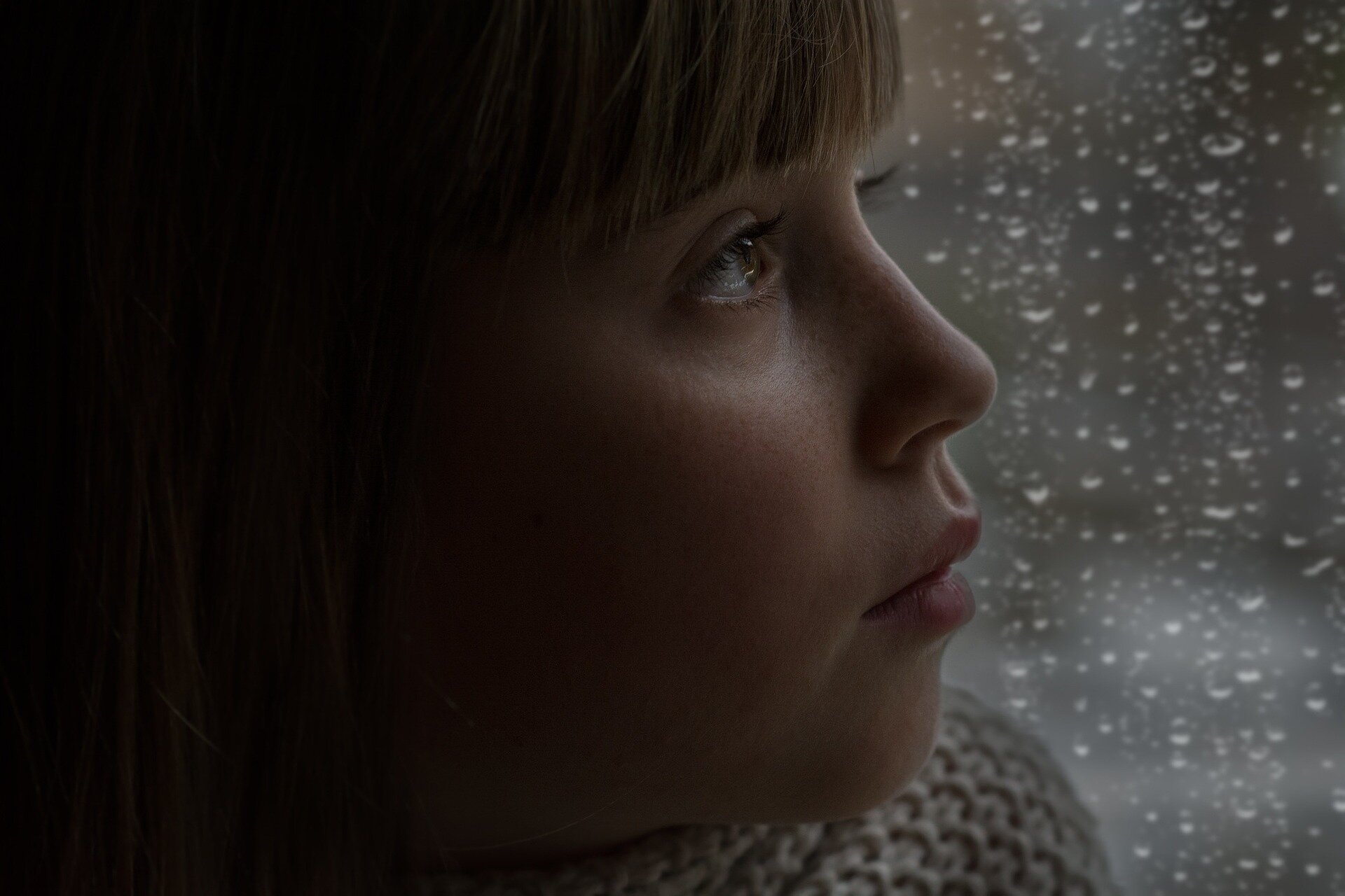 Ilustracja przedstawia dziewczynkę patrzącą przez okno na padający deszcz.