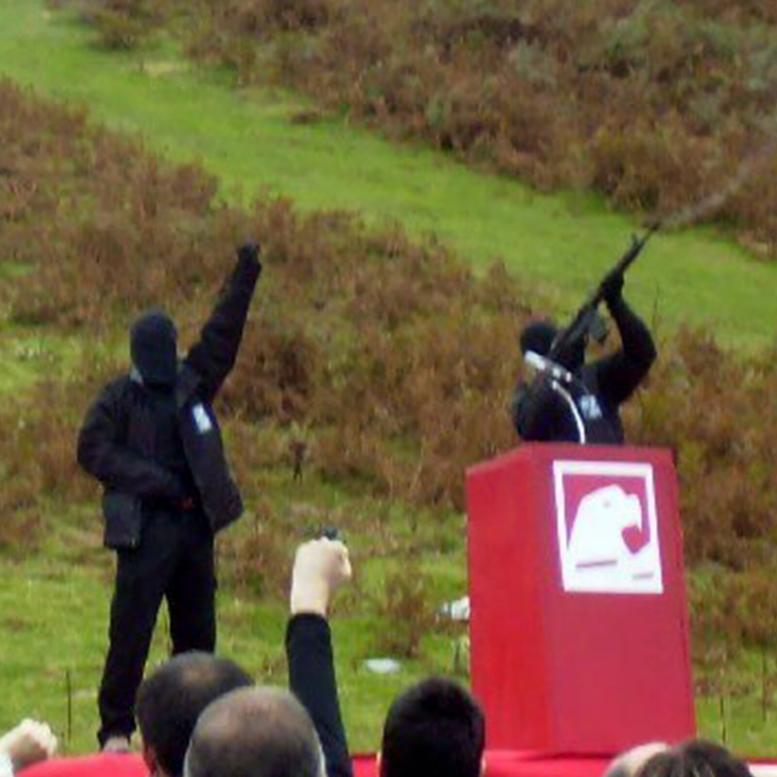 Na zdjęciu widoczni są uzbrojeni członkowie ETA. Są ubrani na czarno oraz mają zasłonięte twarze. Stoją na podwyższeniu. Dwóch z nich kieruje broń w niebo. Trzeci podnosi rękę w górę. Poniżej stoi grupa ludzi. 