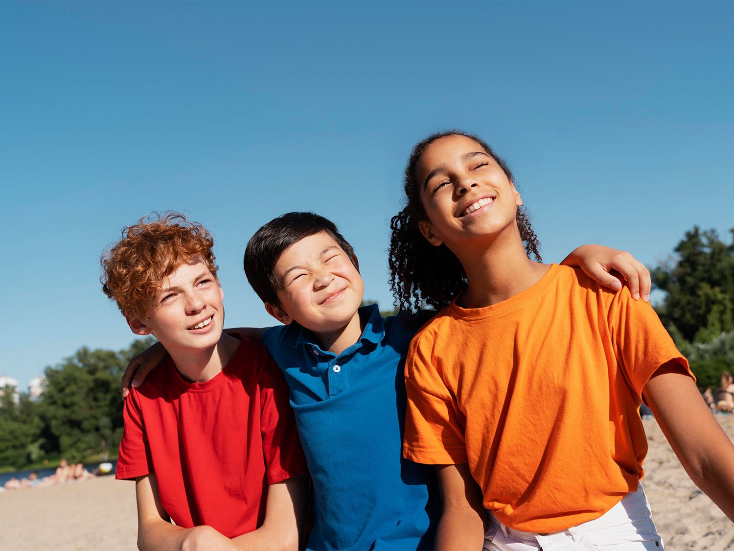Zdjęcie przedstawia tróję stojących, uśmiechniętych dzieci, widocznych do pasa. Dzieci stoją obok siebie, od lewej dwóch chłopców i dziewczynka. Chłopiec stojący pośrodku trzyma rozłożone ręce na ramionach chłopaka i dziewczyny stojących po bokach. Głowy dzieci są uniesione do góry, spoglądają w prawo, mają lekko zmrużone oczy. W tle widoczny pas zielonych drzew.   