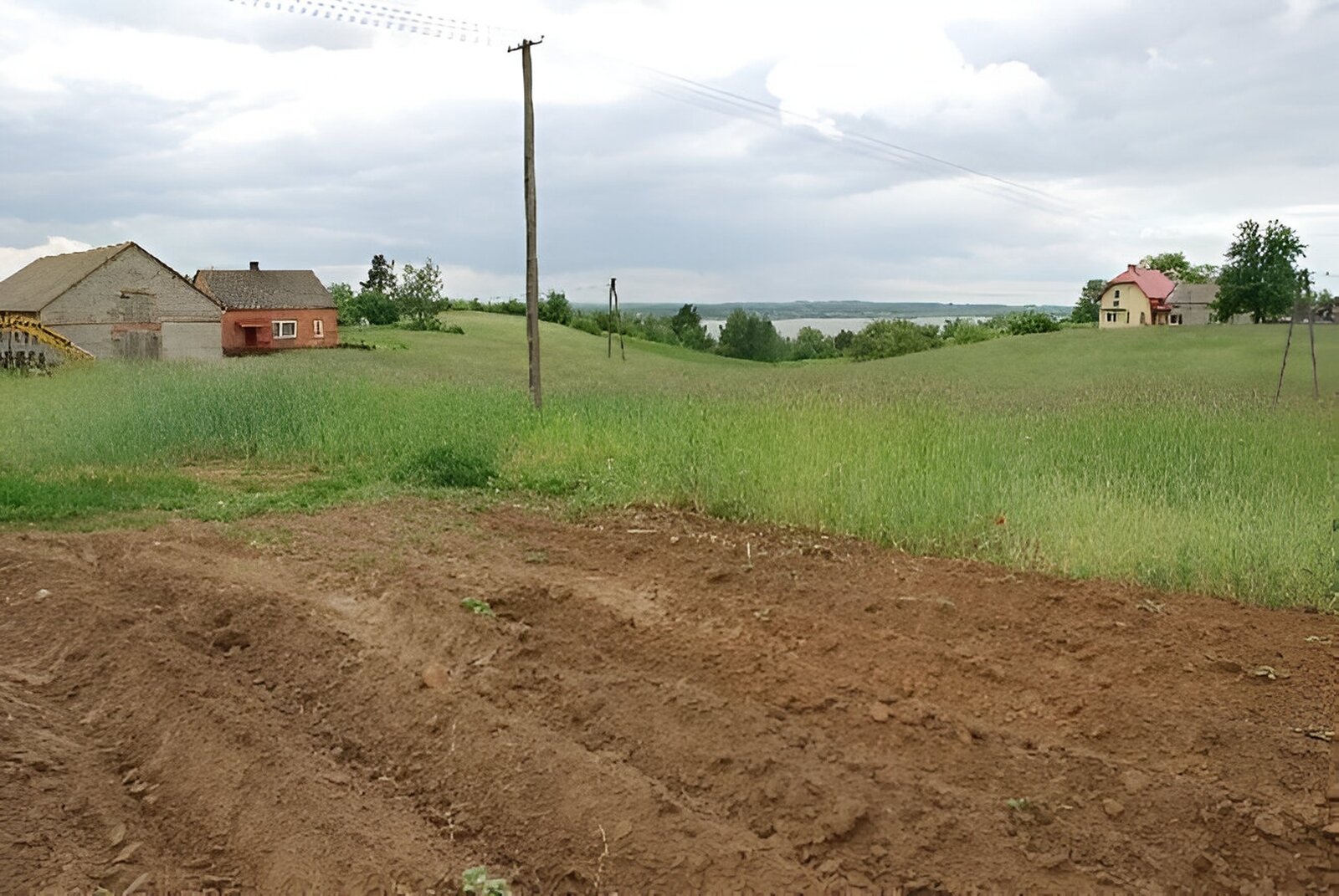 Zdjęcie przedstawia pole częściowo zaorane. W oddali pagórki, wokół nich łąka i domy. 