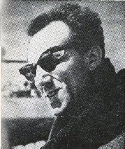 Zdjęcie przedstawia lewy profil dojrzałego mężczyzny. Twarz mężczyzny jest pociągła, włosy krótkie. Mężczyzna ma ciemne okulary, uśmiecha się lekko.