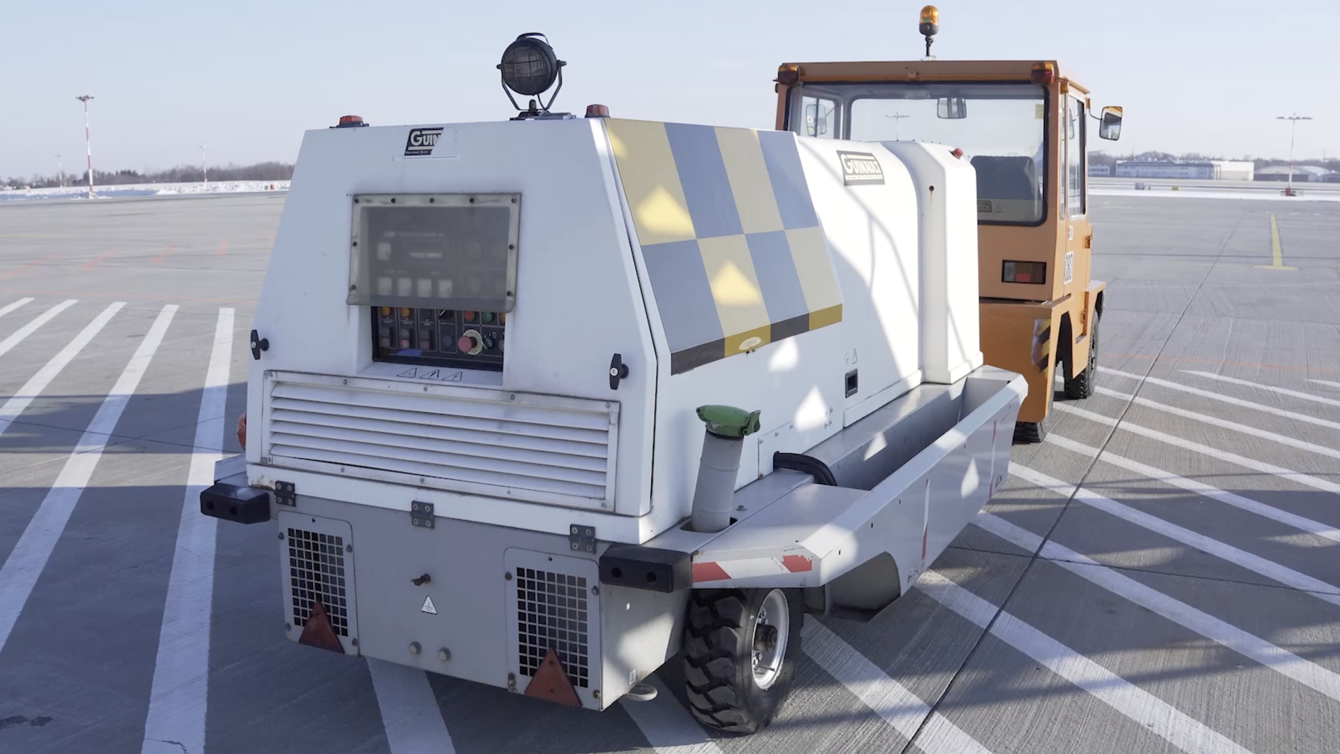 Zdjęcie przedstawia G P U, czyli agregat prądotwórczy zasilany paliwem. Jest to prostopadłościenne urządzenie na kołach podłączone do niewielkiego ciągnika. Na pionowej tylnej ścianie urządzenia znajduje się panel sterowania. Na nim znajduje się wiele przycisków.