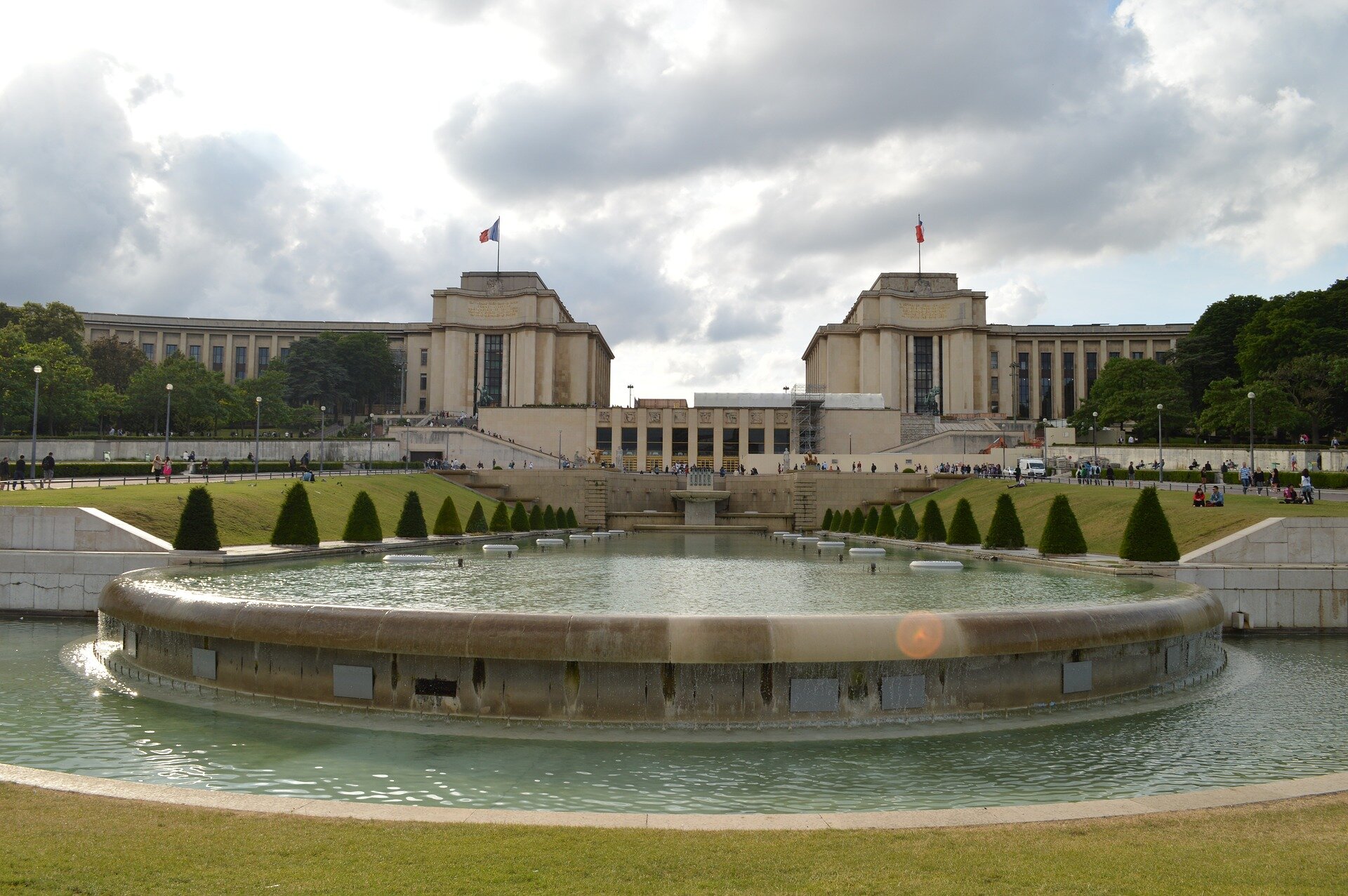 Fotografia przedstawia fontannę na placu Trocadero. Fontana ma dwa poziomy i jest podłużna. Po bokach fontanny rosną małe drzewka. Całość otoczona przystrzyżonym trawnikiem. Za fontanną, nieco powyżej widoczna jest centralna część pałacu. Niebo jest zachmurzone.