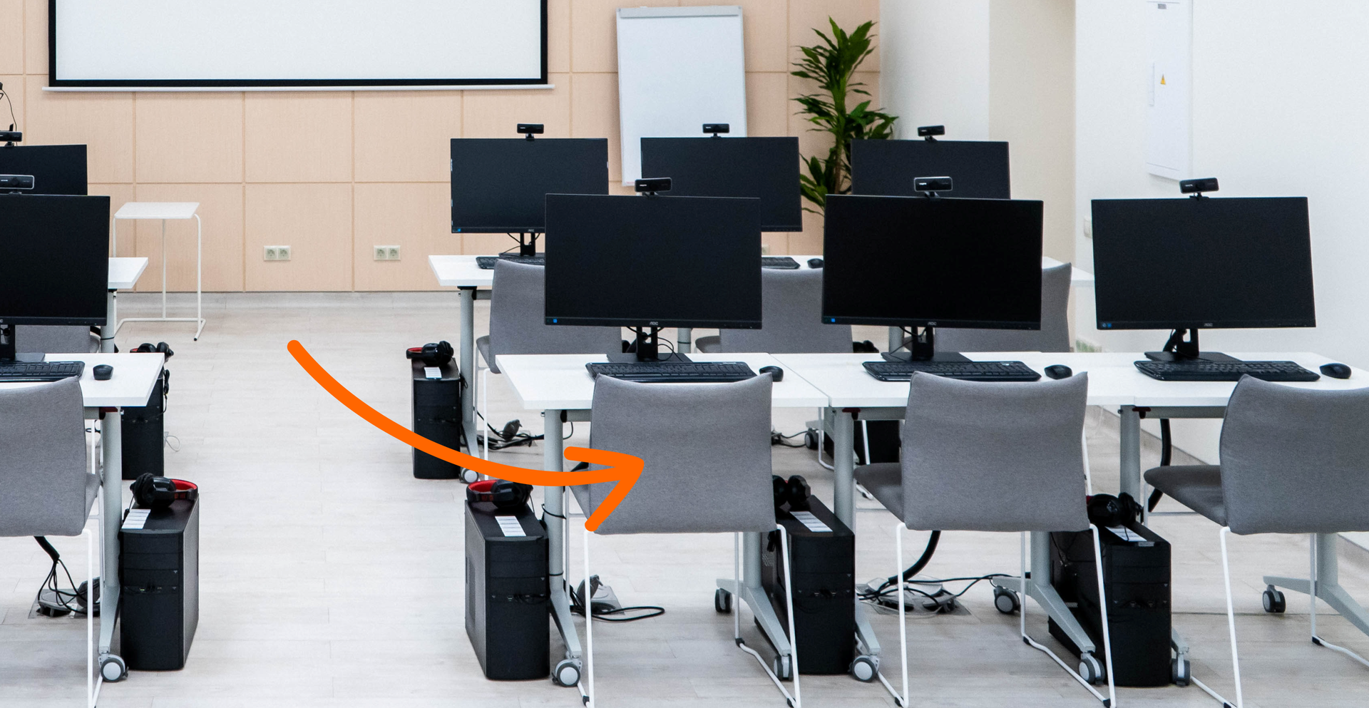 Zdjęcie przedstawia stanowiska komputerowe ustawione w rzędach. Na każdym stoliku znajduje się monitor, obok stoi stacja komputera. Przy każdym stoliku znajduje się krzesło. Na ilustracji widoczna jest strzałka skierowana na jedno z krzeseł.