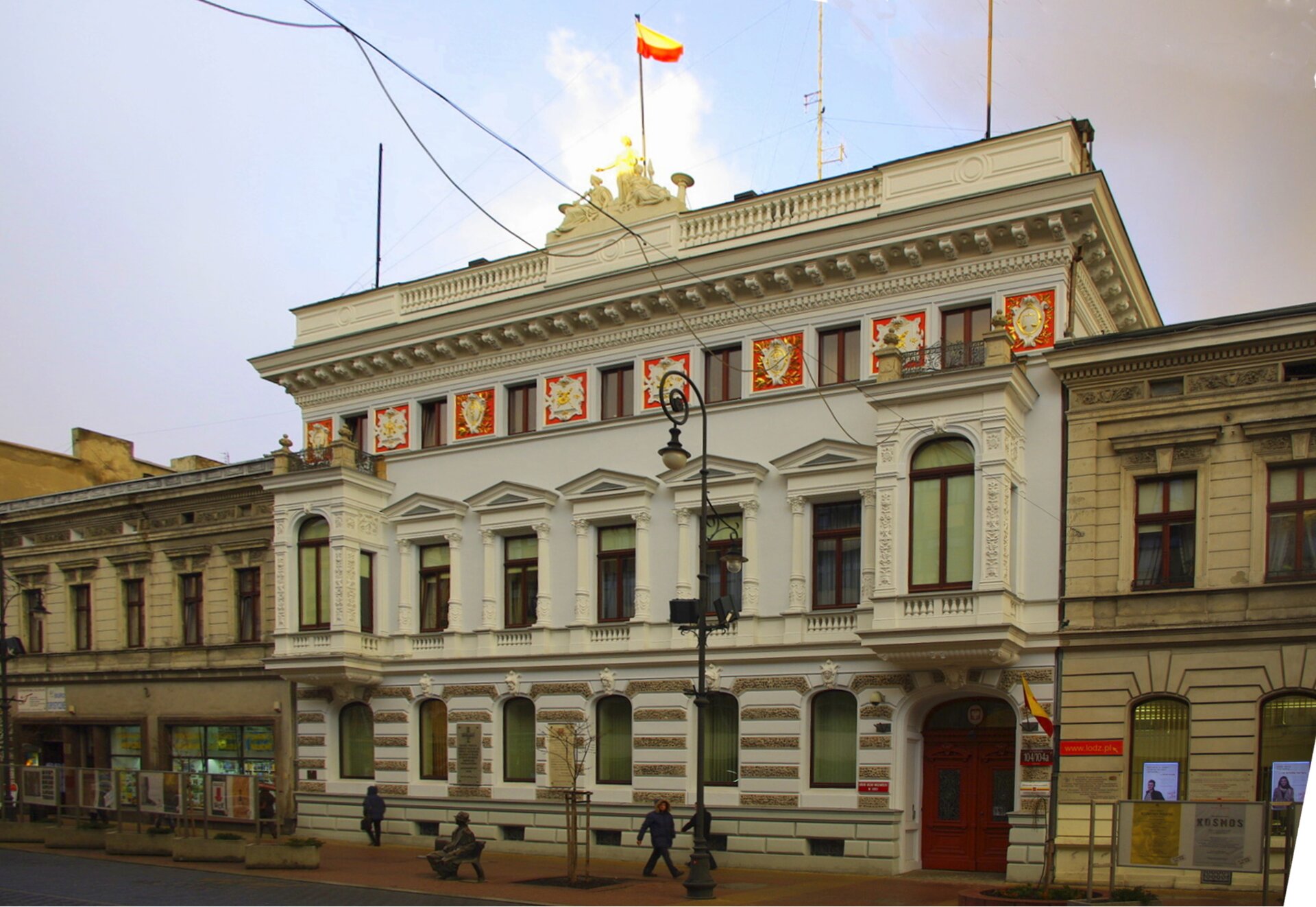 Ilustracja o kształcie poziomego prostokąta przedstawia zdjęcie Pałacu w Łodzi autorstwa Hilarego Majewskiego. Zdjęcie ukazuje jasny budynek, przed którym widać przechodniów. Obiekt jest trójkondygnacyjny, trzecia kondygnacja jest niższa od pozostałych, dolnych. Dolna natomiast dekorowana jest rustyką.   Budowlę zamyka balustrada z dekoracją rzeźbiarską. 