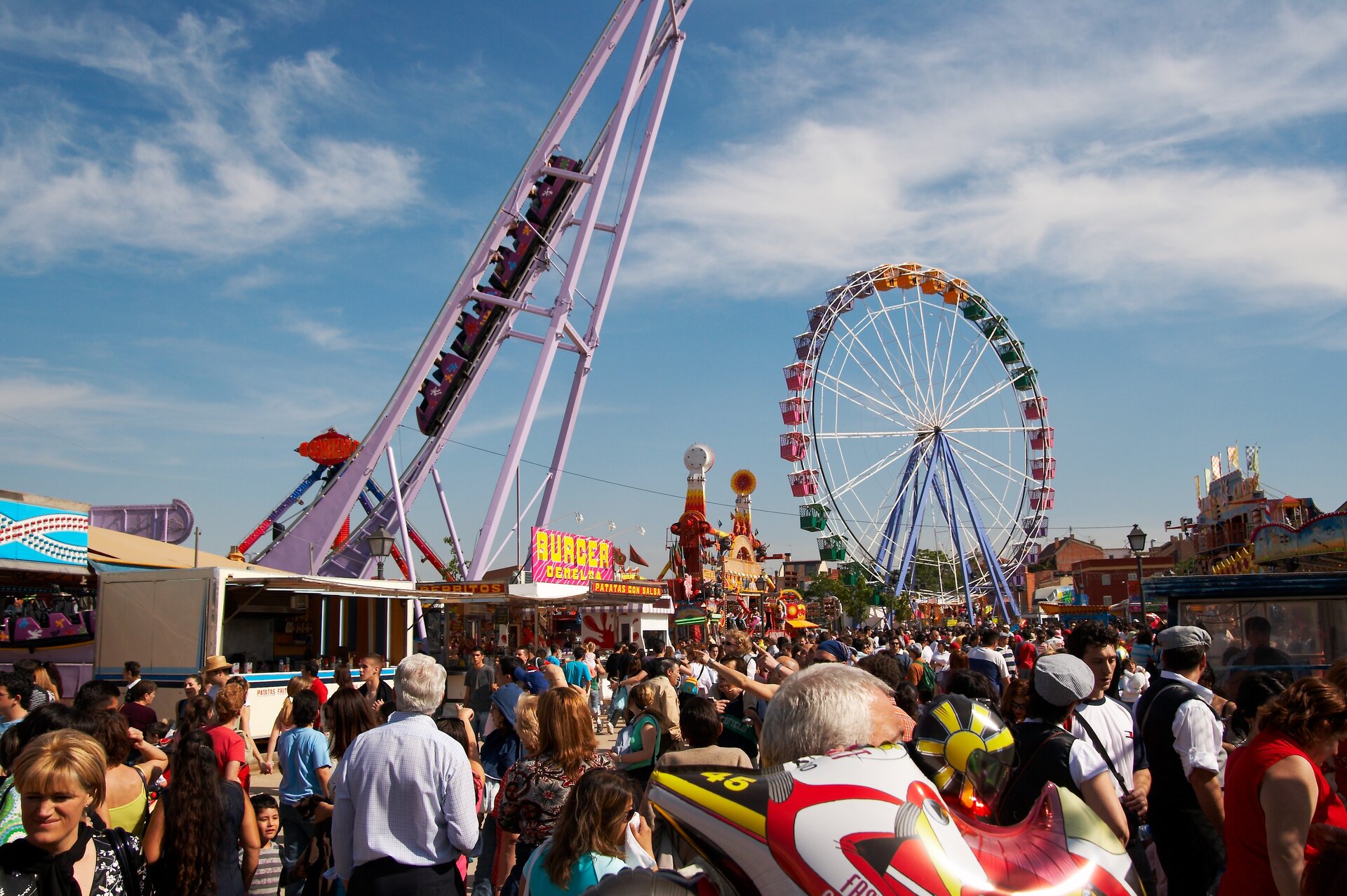 Zdjęcie przedstawiajace fragment wesołego misteczka. Na pierwszym planie tłum ludzi.  Na drugim planie kolejka górska i diabelski młyn.