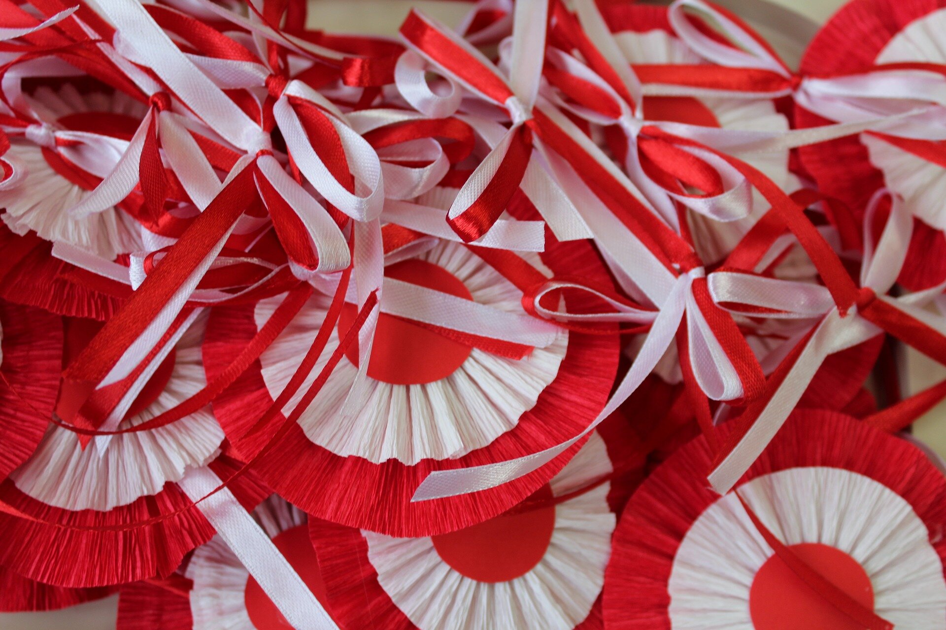 Zdjęcie przedstawia biało‑czerwone kokardy narodowe oraz kokardki wykonane ze wstążek również w kolorze biało‑czerwonym.