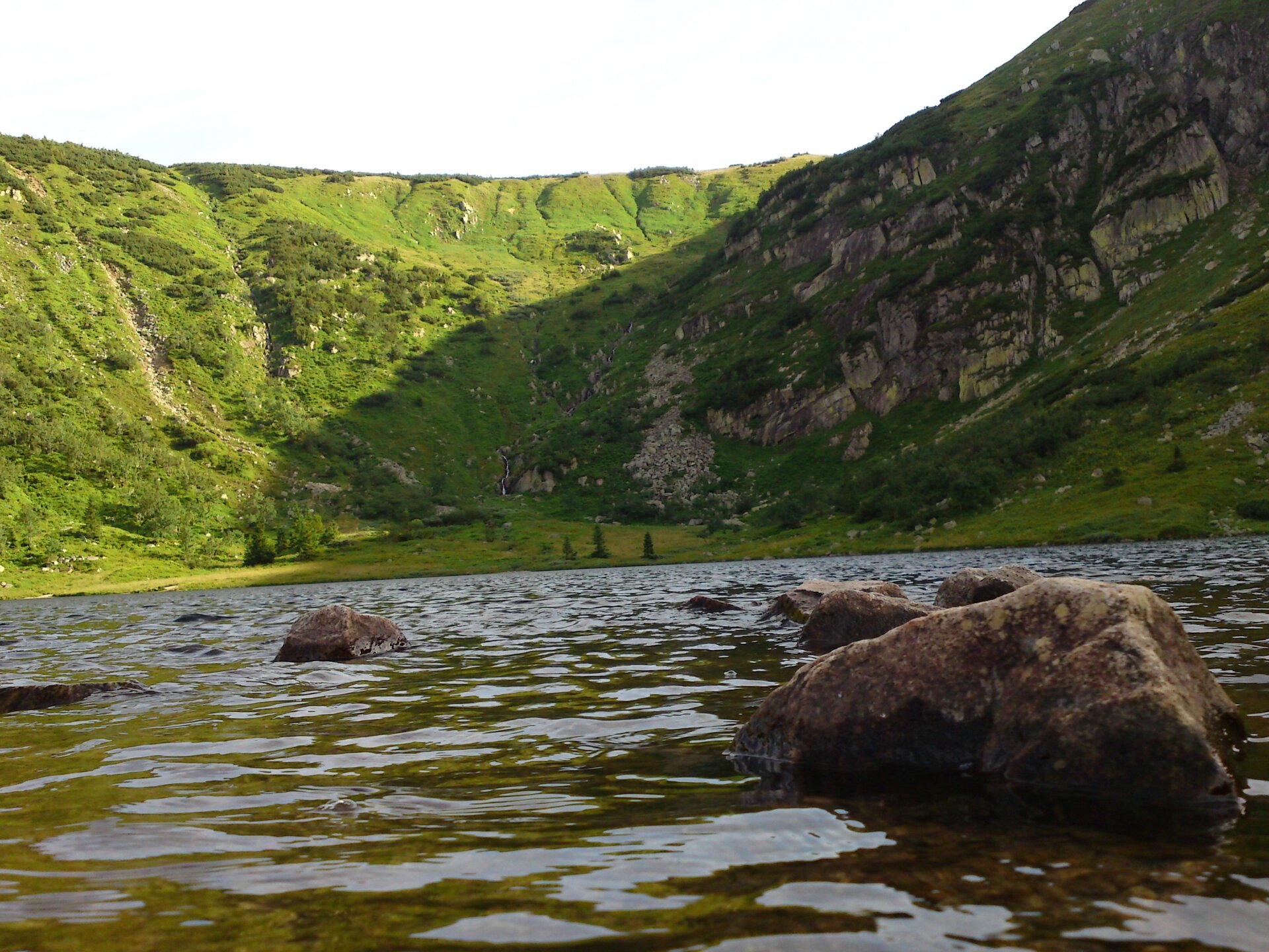 Fotografia jeziora otoczonego wzniesieniami pokrytymi trawami, krzewami, gdzieniegdzie małymi drzewami. Na pierwszym planie z wody wystają pojedyncze głazy.