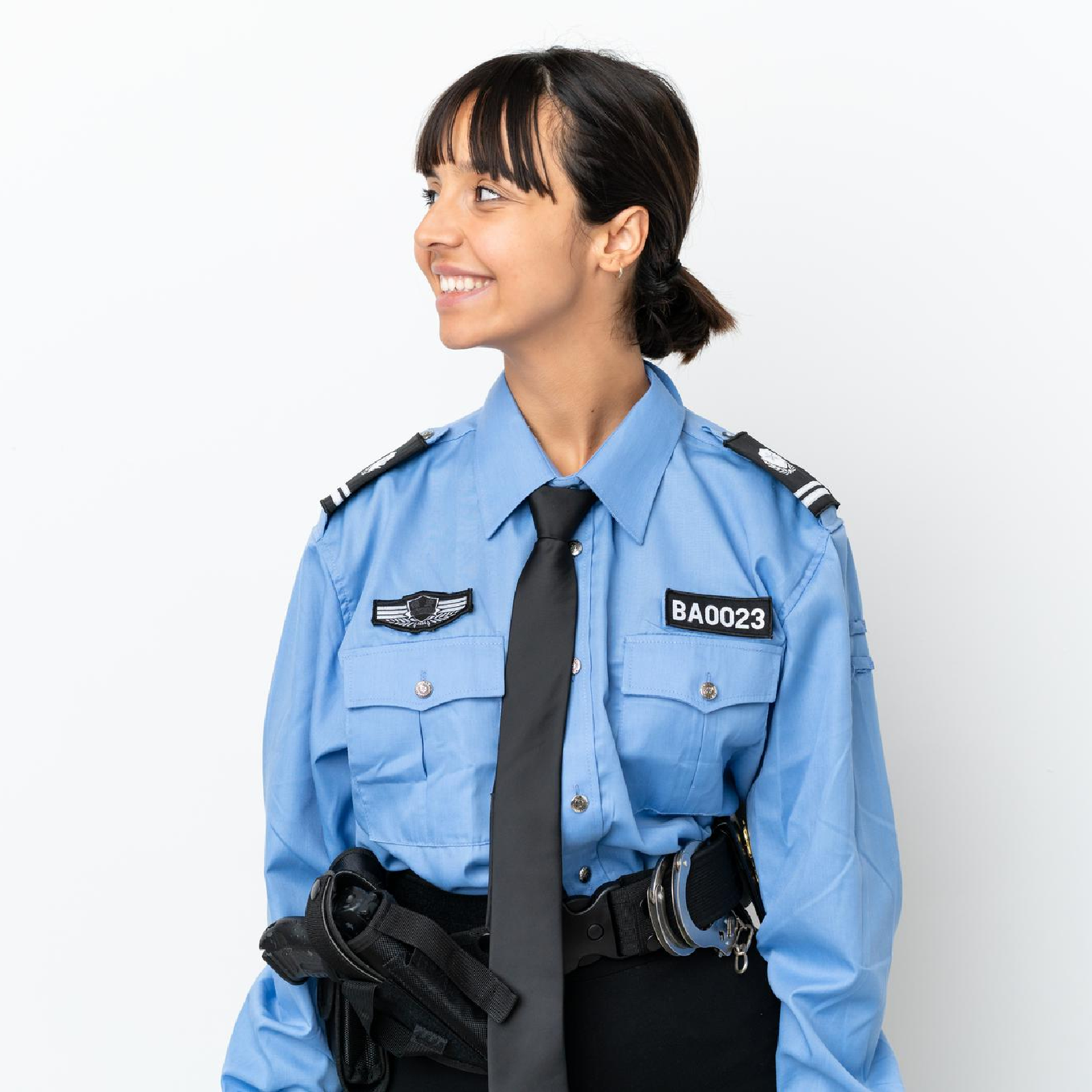 Ilustracja przedstawia kobietę w stroju policjantki. Ma niebieską koszulę z długim rękawem, oraz pagonami na ramionach. Pod szyją, zawiązany ma czarny krawat. Na prawej piersi, przyszyty ma emblemat. Na lewej piersi, przyszyty ma numer: B A 0023. Przy pasie, widoczne są kajdanki oraz kabura na gaz obezwładniający.