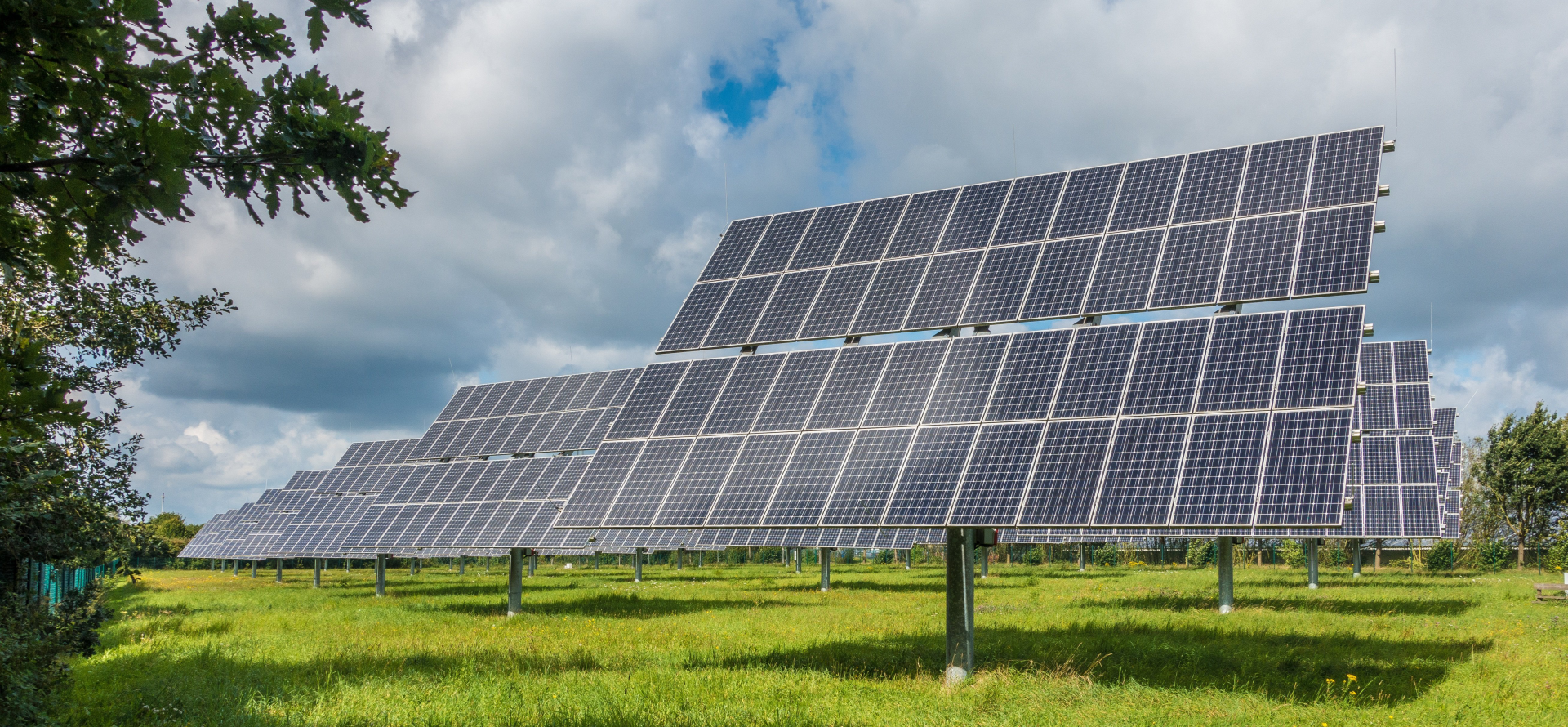Zdjęcie przedstawia rzędy paneli fotowoltaicznych ustawionych na łące. Panele mają kształt szarych prostokątów z naniesionymi białymi cienkimi kratkami. Prostokąty tworzą całe połacie prostokątów. Jasnozielona trawa krótko przycięta. Niebo zachmurzone.