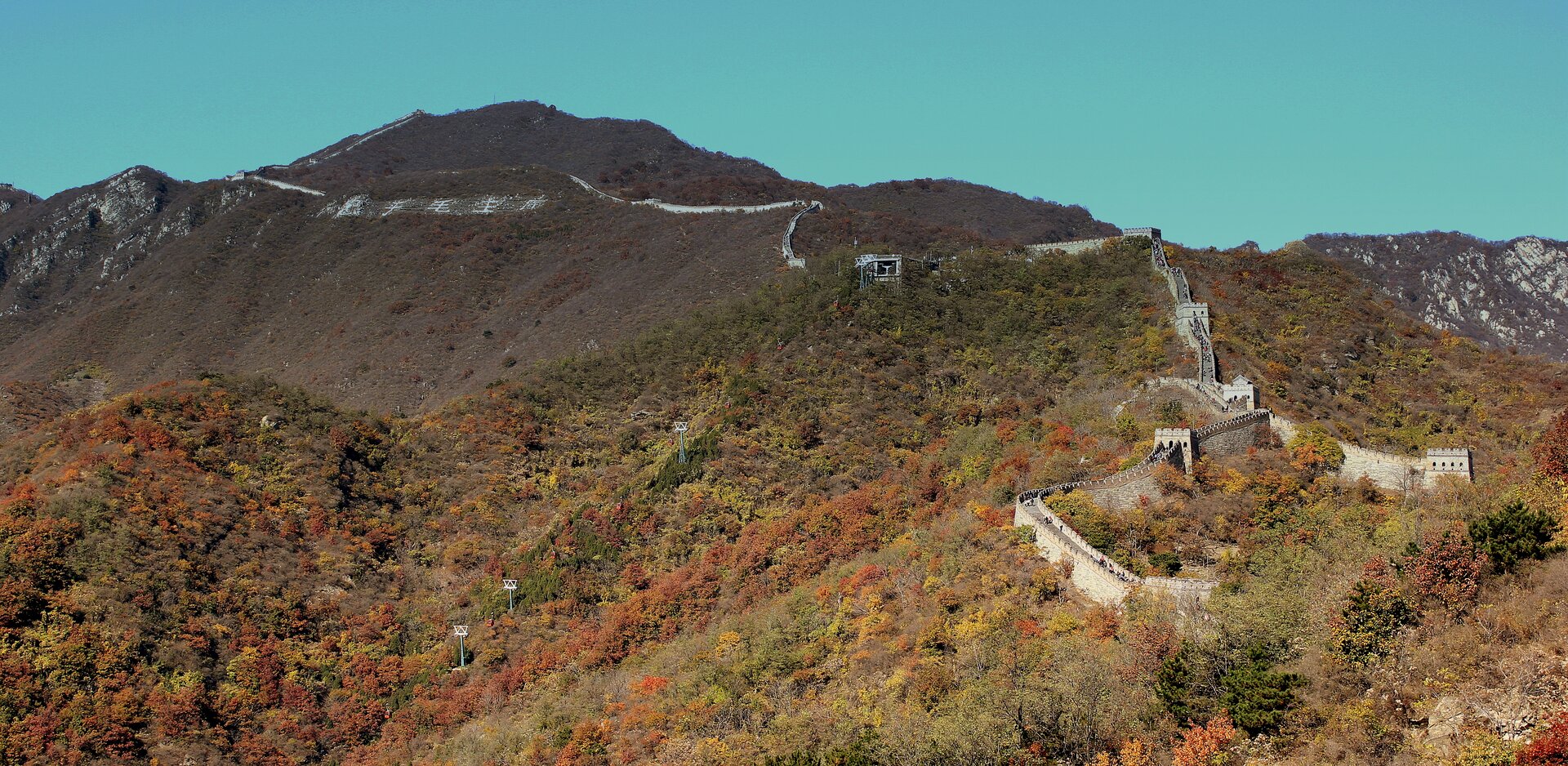 Na zdjęciu góry. Stoki gór widocznych na pierwszym planie pokrywa roślinność – las liściasty podczas jesieni. Kolory zielone, żółte, czerwone i brązowe. Wyższe partie gór słabiej pokryte roślinnością. Wzdłuż górskich szczytów wysoki i szeroki mur. Na murze droga, po której spacerują turyści. Co kilkaset metrów na murze jest wieża obserwacyjna.