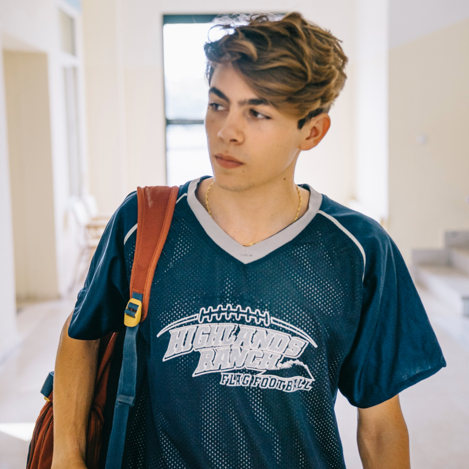 Zdjęcie przedstawia młodego chłopaka, od pasa w górę. Chłopak ma jasne, krótkie włosy, głowę zwróconą w lewo, zamyślony wyraz twarzy. Ubrany jest w sportową koszulkę z krótkim rękawem, w kolorze niebieskim, z napisem w języku angielskim: Highlands Ranch Flag Football. Na prawym ramieniu chłopak ma przerzucony plecak. W tle widoczny jest korytarz i duże okno.
