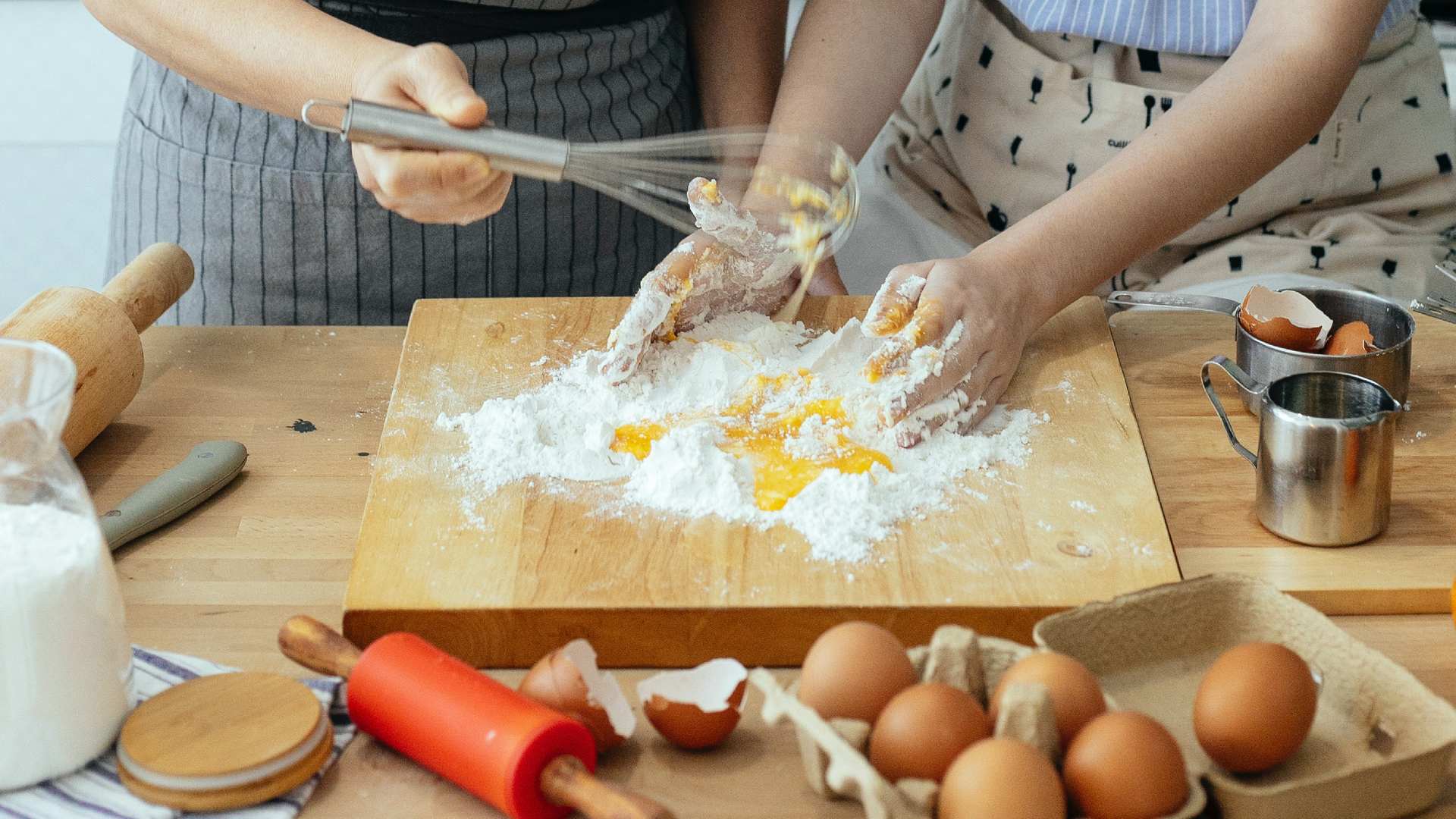 Zdjęcie przedstawia przygotowywanie ciasta. Dwie osoby stojące przy blacie, na którym leży stolnica. Na stolnicy znajdują się mąka i rozbite jajka. Osoba z lewej strony miesza jajka trzepaczką, natomiast osoba z prawej strony rękoma ugniata składniki. Na blacie znajdują się również rondelek ze skorupkami jajek, drewniany wałek do ciasta, metalowy dzbanek,  mniejszy silikonowy wałek oraz jajka w wytłaczance.