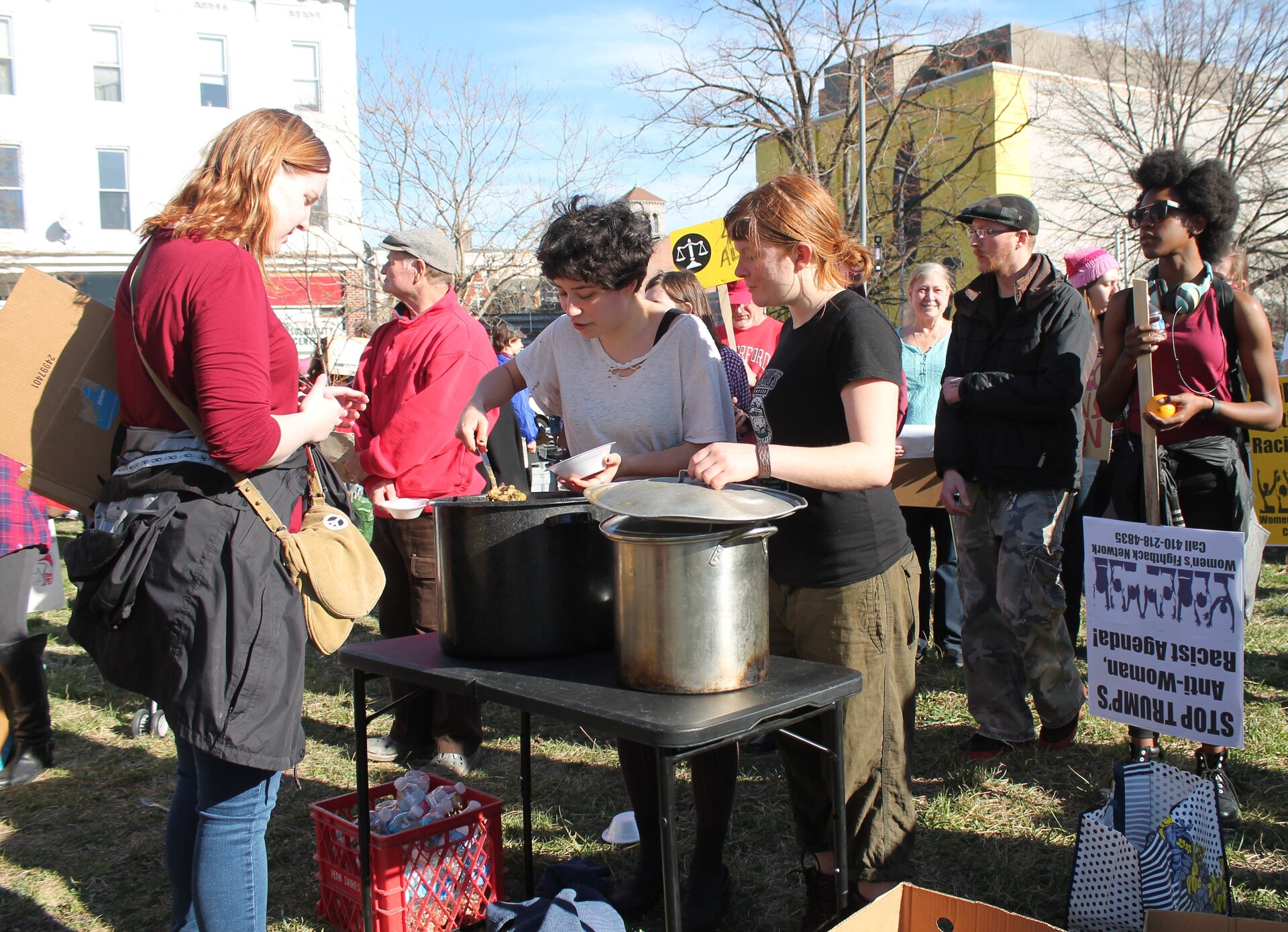 Zdjęcie przedstawia grupę ludzi stojących na trawie w mieście. Na pierwszym planie dwie kobiety nabierają do miski jedzenie z dużych garnków stojących na stoliku. Na jedzenie czeka młoda kobieta. Za nimi ludzie z transparentami. Na jednym z nich, trzymanym przez kobietę, napis: "Stop Trump's Anti-Woman. Racist Agenda".  