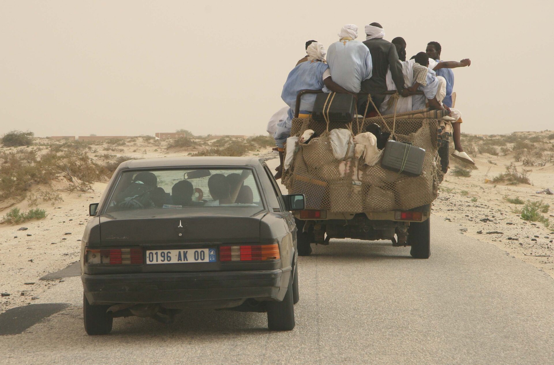 Na zdjęciu dwa samochody (osobowy i pick‑up) na drodze w piaszczystym terenie. Samochody są zatłoczone. Na pick‑upie jest mnóstwo bagaży i pasażerów.