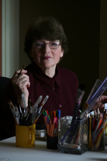 Kliknij, aby powiększyć. Kolorowe zdjęcie przedstawia pisarkę siedzącą za biurkiem, na którym stoją pojemniki z długopisami, pisakami, kredkami, pędzelkami i linijkami. Kobieta ma krótkie ciemne włosy i okulary.