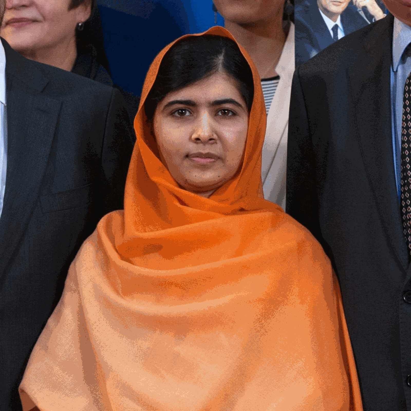 Zdjęcie przedstawia Pakistankę, w ujęciu od pasa w górę, stojącą pomiędzy ludźmi w garniturach i żakietach. Kobieta ma ciemne włosy schowane pod pomarańczową chustą okrywającą jej głowę, ramiona i klatkę piersiową. Kobieta patrzy prosto w obiektyw.