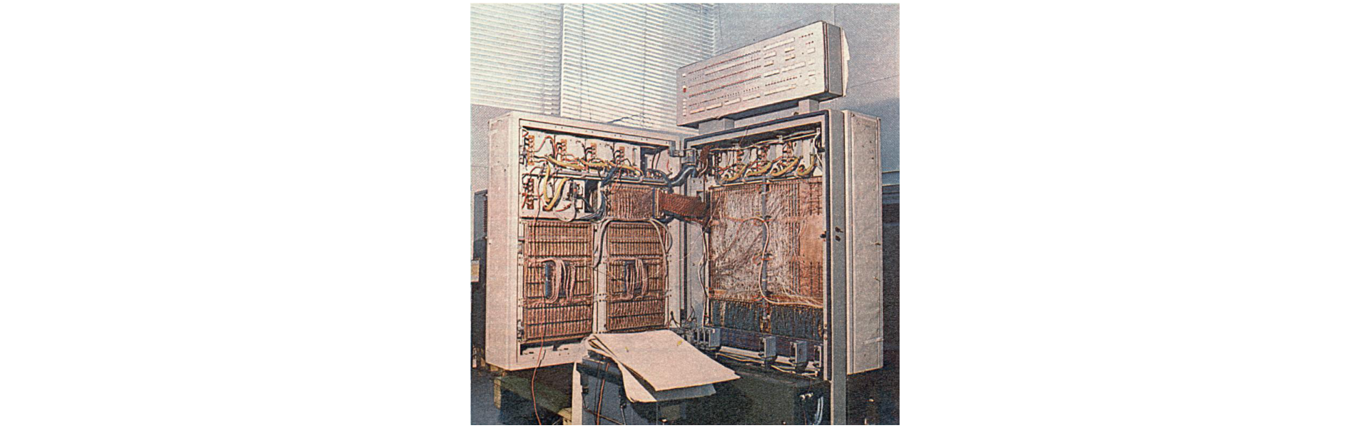 Na zdjęciu widoczny jest pokój, w oknach wiszą poziome żaluzje. Na pierwszym planie widać jeden z pierwszych komputerów Odra. Jest to duża szafa, w której znajdują się układy scalone łączone przewodami. Przed komputerem leży taśma perforowana - arkusze papieru.